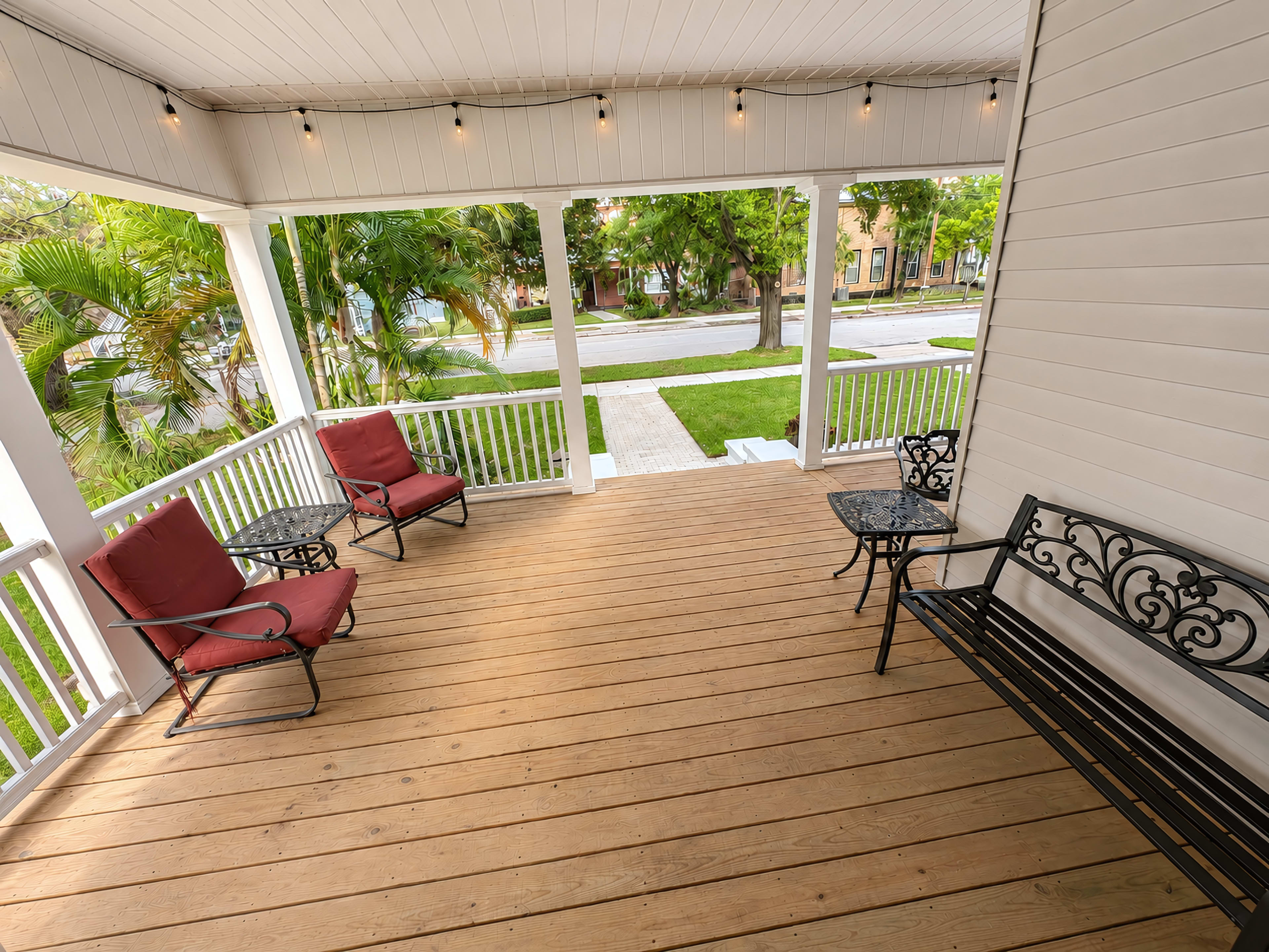 The image shows a spacious porch with wooden flooring, two red lounge chairs, a small round table, and a black metal bench, surrounded by green trees and a pathway.