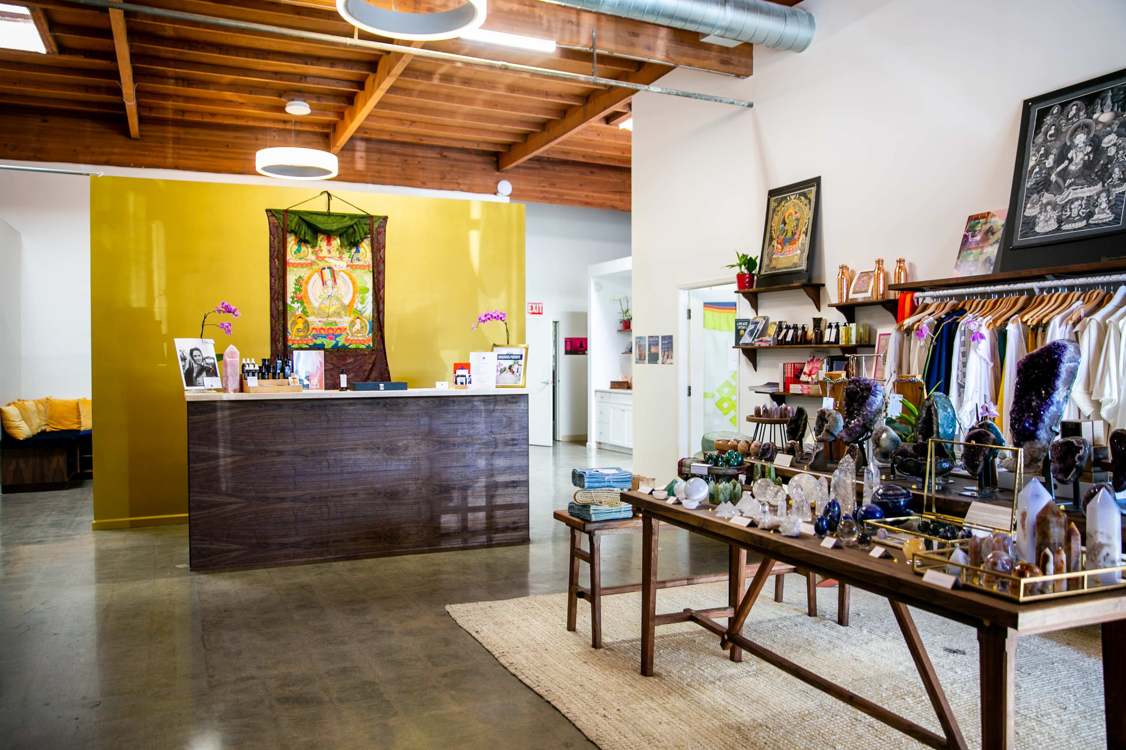 The interior of a wellness shop features a wooden reception desk against a bright yellow wall, surrounded by various crystals and wellness products displayed on tables and shelves.
