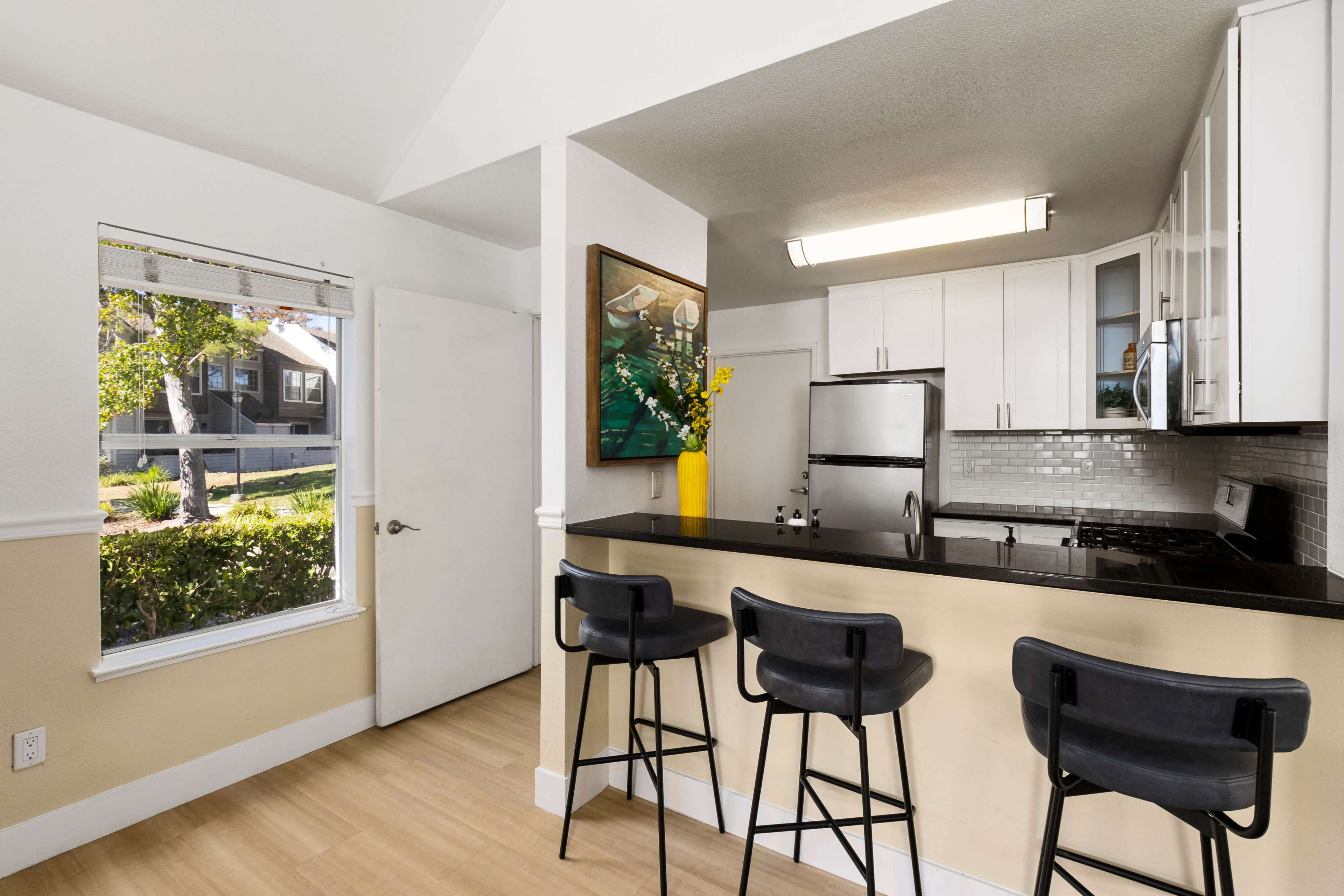 A modern kitchen features white cabinets, stainless steel appliances, and a dark countertop with four bar stools, adjacent to a large window providing natural light.