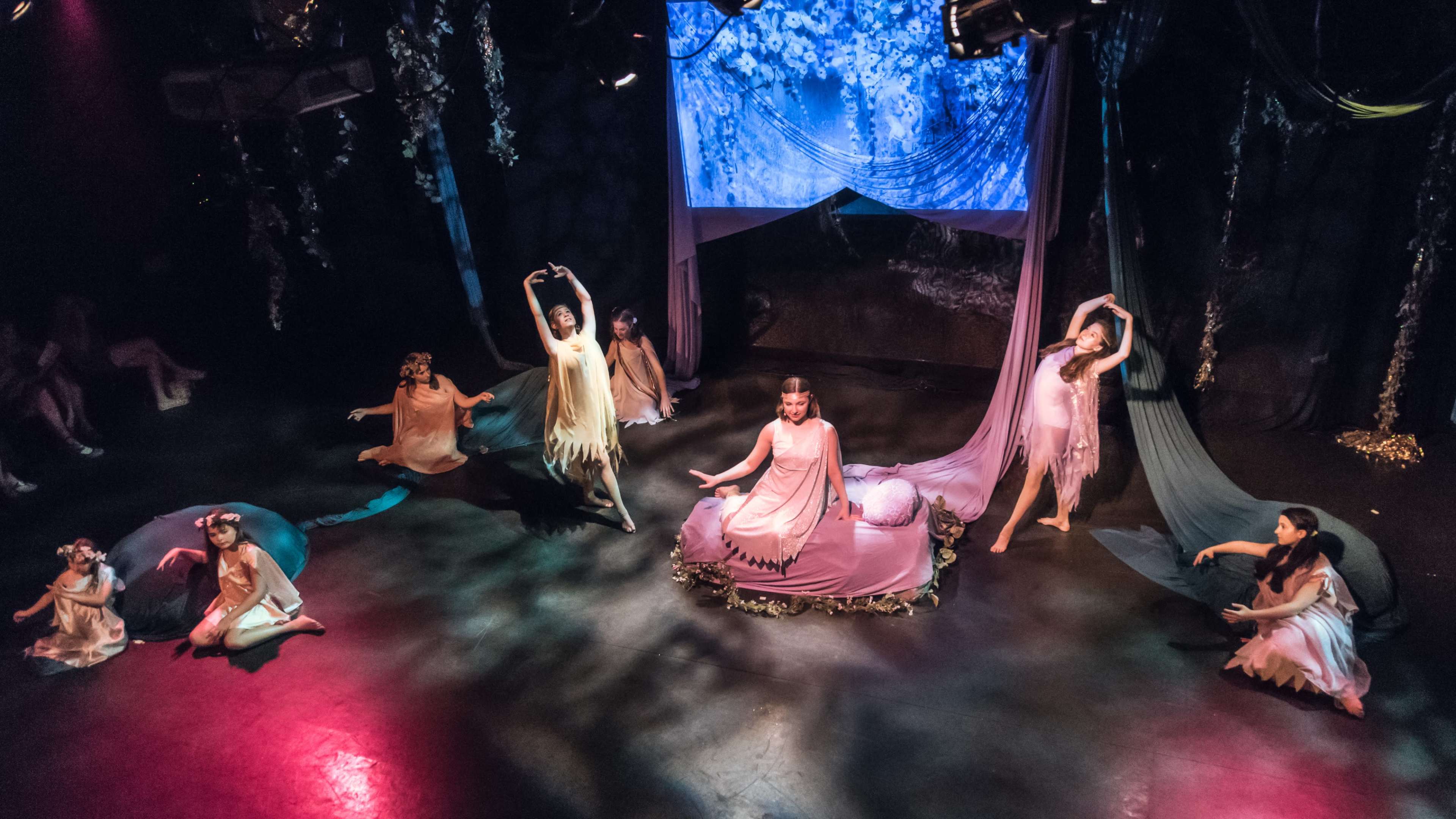 A theatrical performance with several dancers dressed in flowing costumes, performing around a central figure on a stage adorned with fabric and soft lighting.