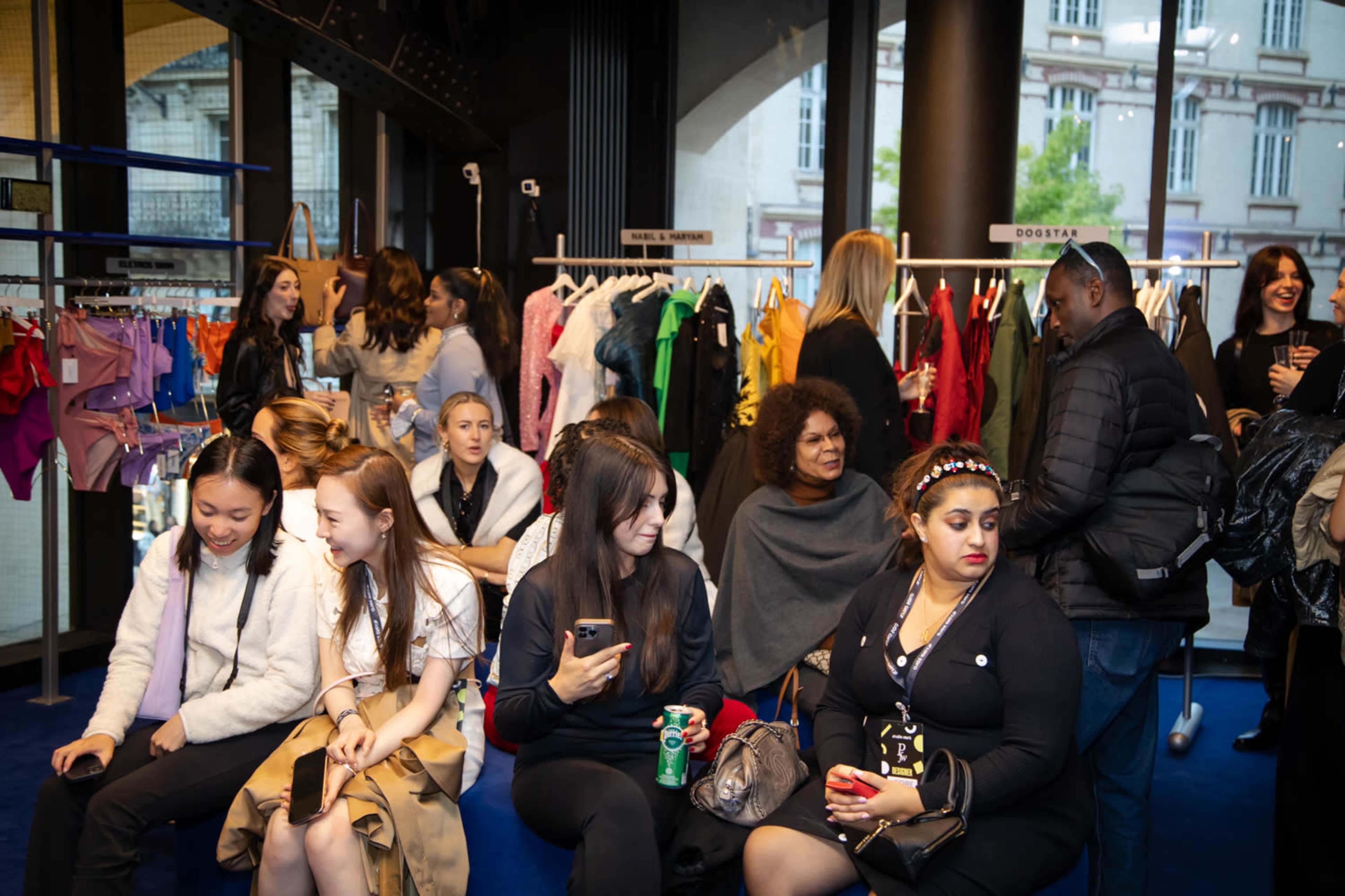 A diverse group of individuals socializes in a modern clothing store with racks of colorful garments in the background.
