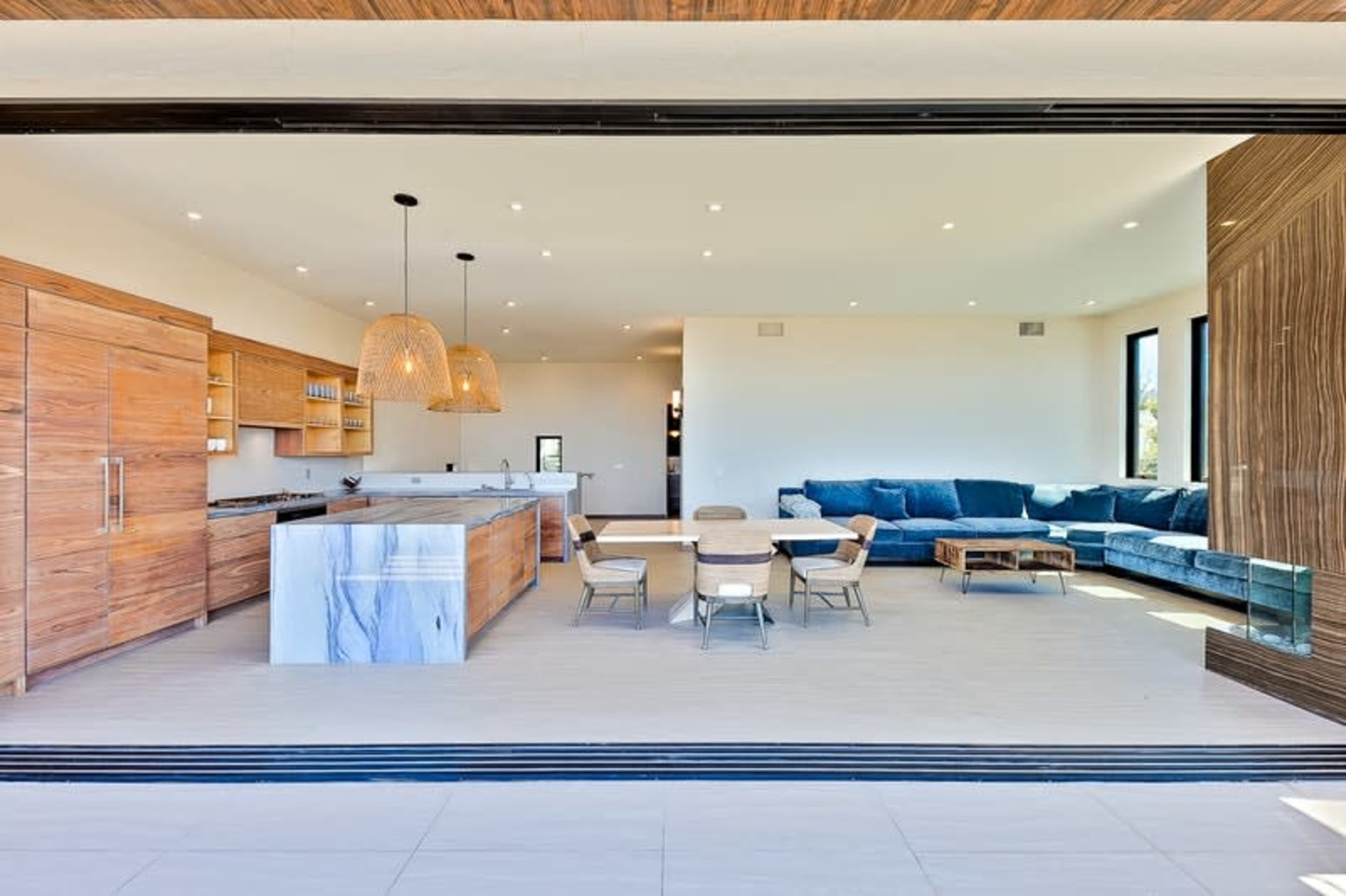 A modern open-concept kitchen and living area features wood cabinetry, an island with a marble countertop, and a sectional sofa against large windows.