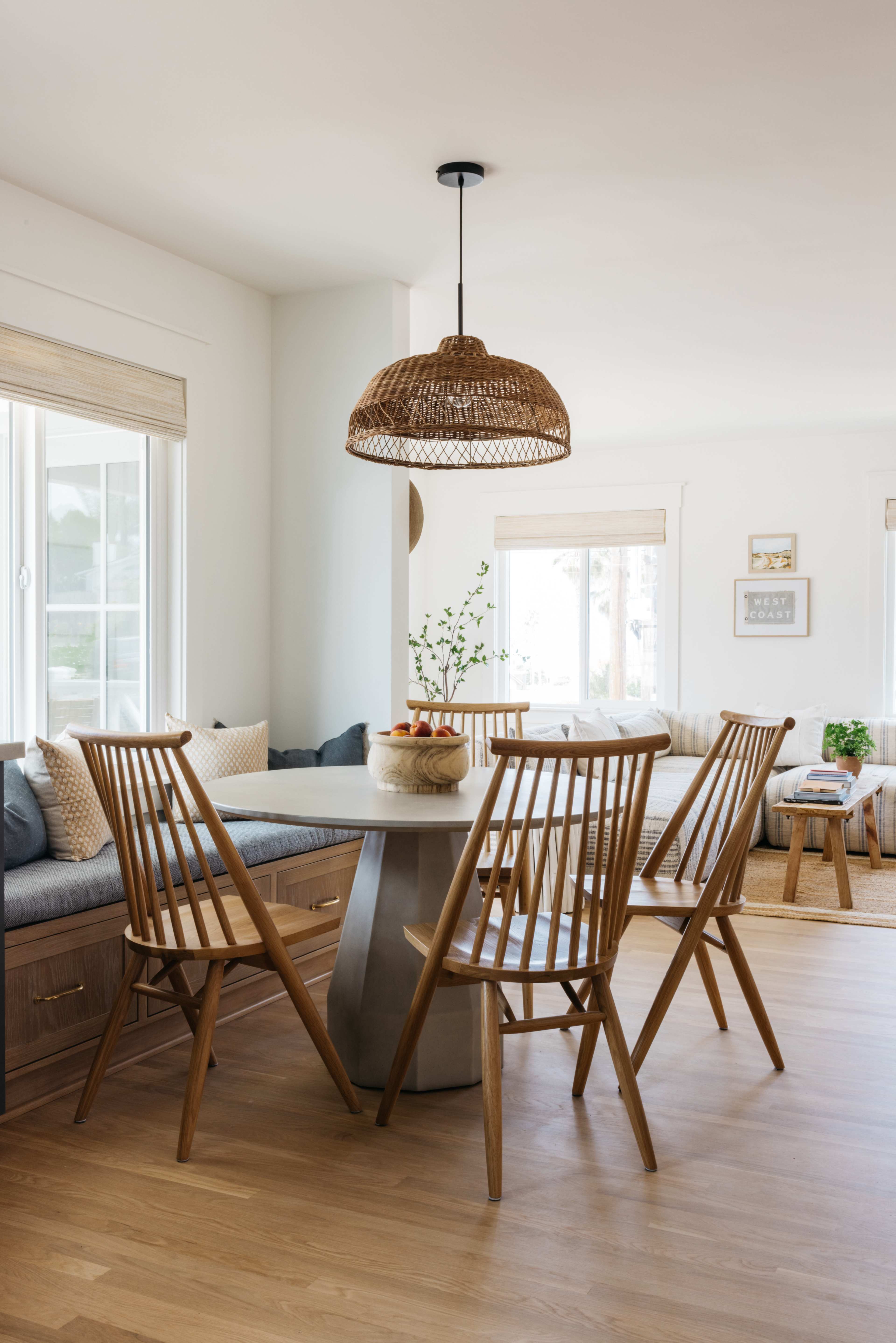 A dining area features a round table surrounded by wooden chairs, with a woven pendant light overhead and large windows allowing natural light.