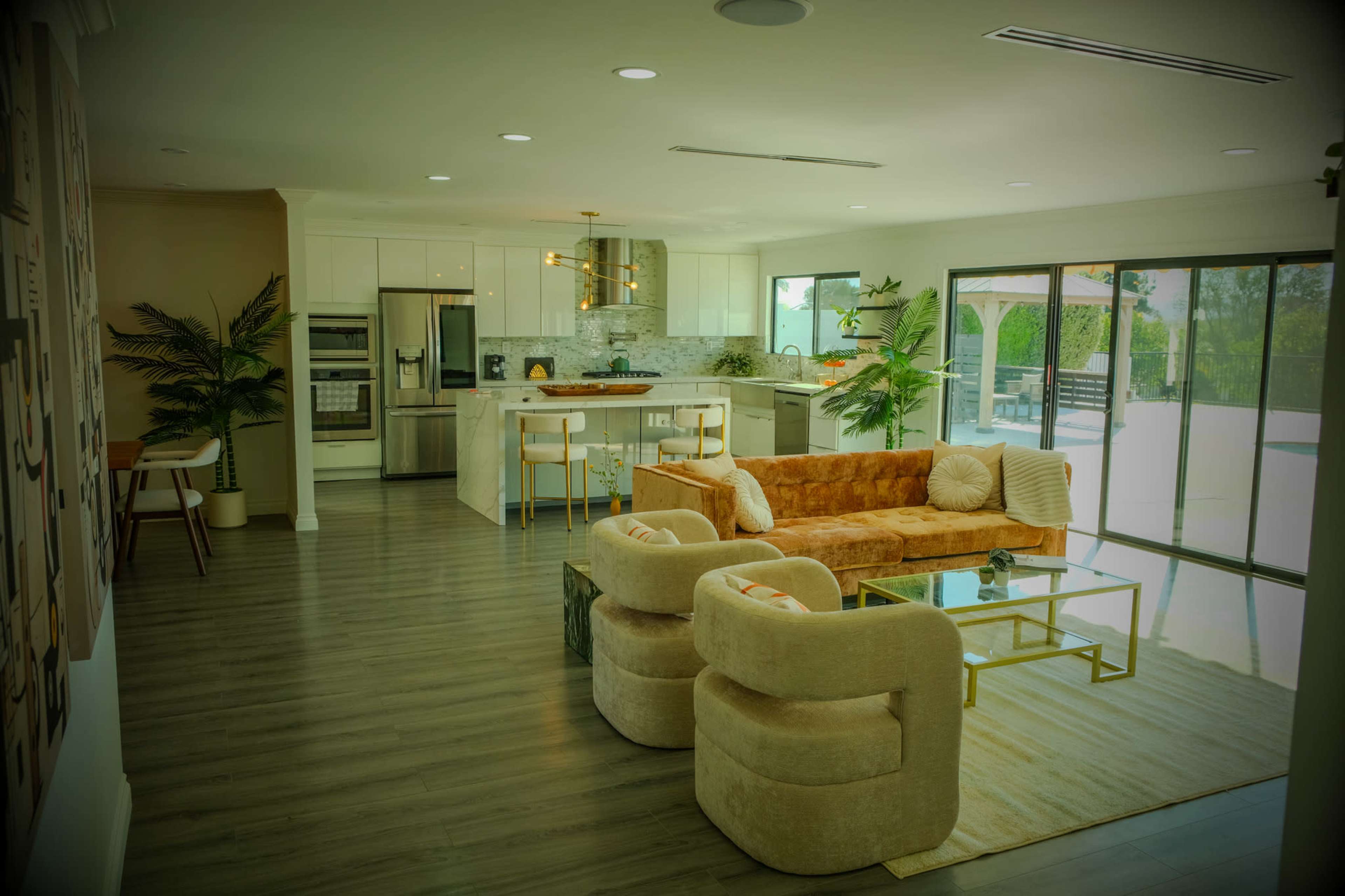 The image shows a modern living space featuring a cozy seating area with an orange sofa and two white armchairs, adjacent to a kitchen equipped with stainless steel appliances and a large window overlooking a patio.