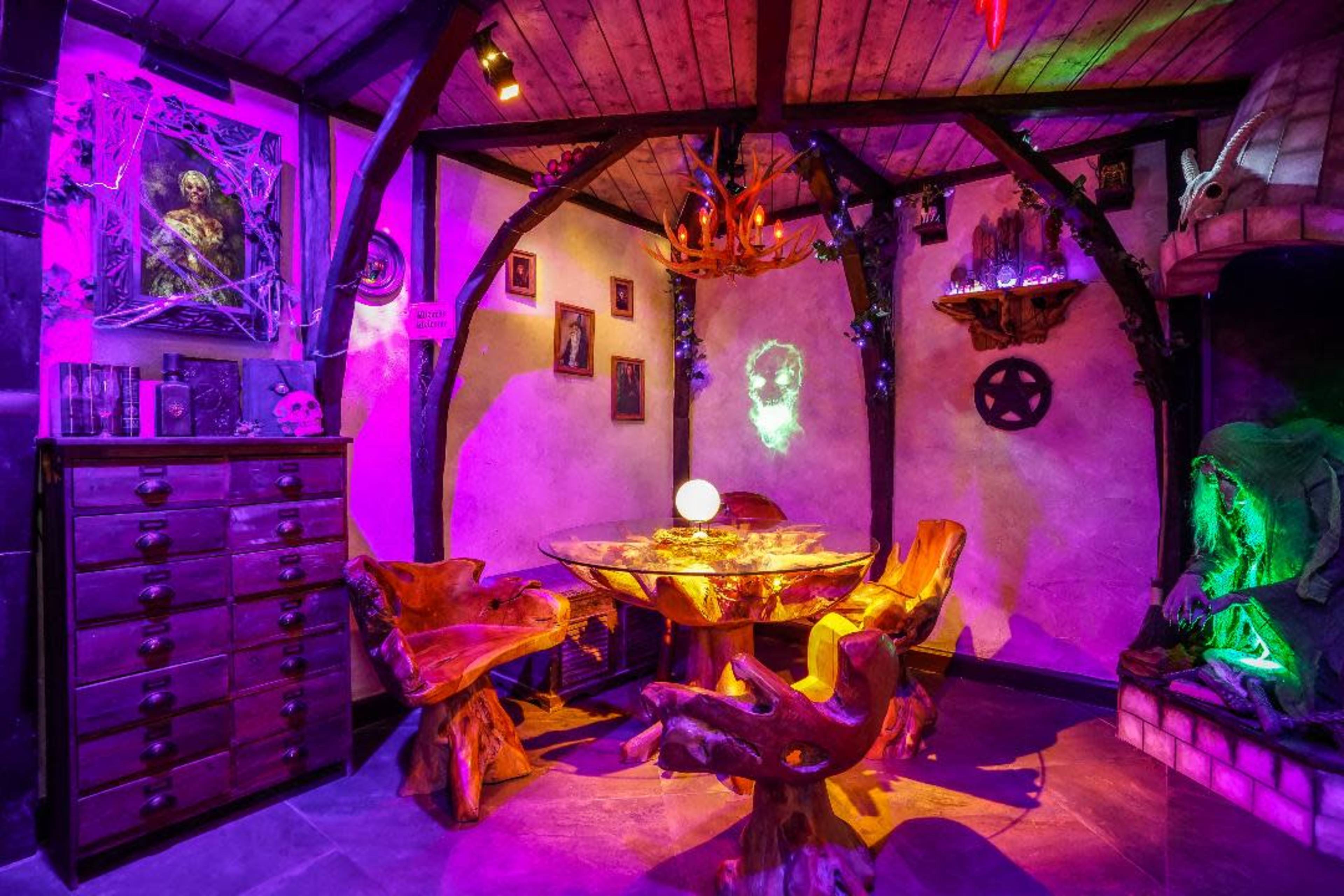 A dimly lit room features a circular table surrounded by unique wooden chairs, with mystical decor and glowing elements on the walls.