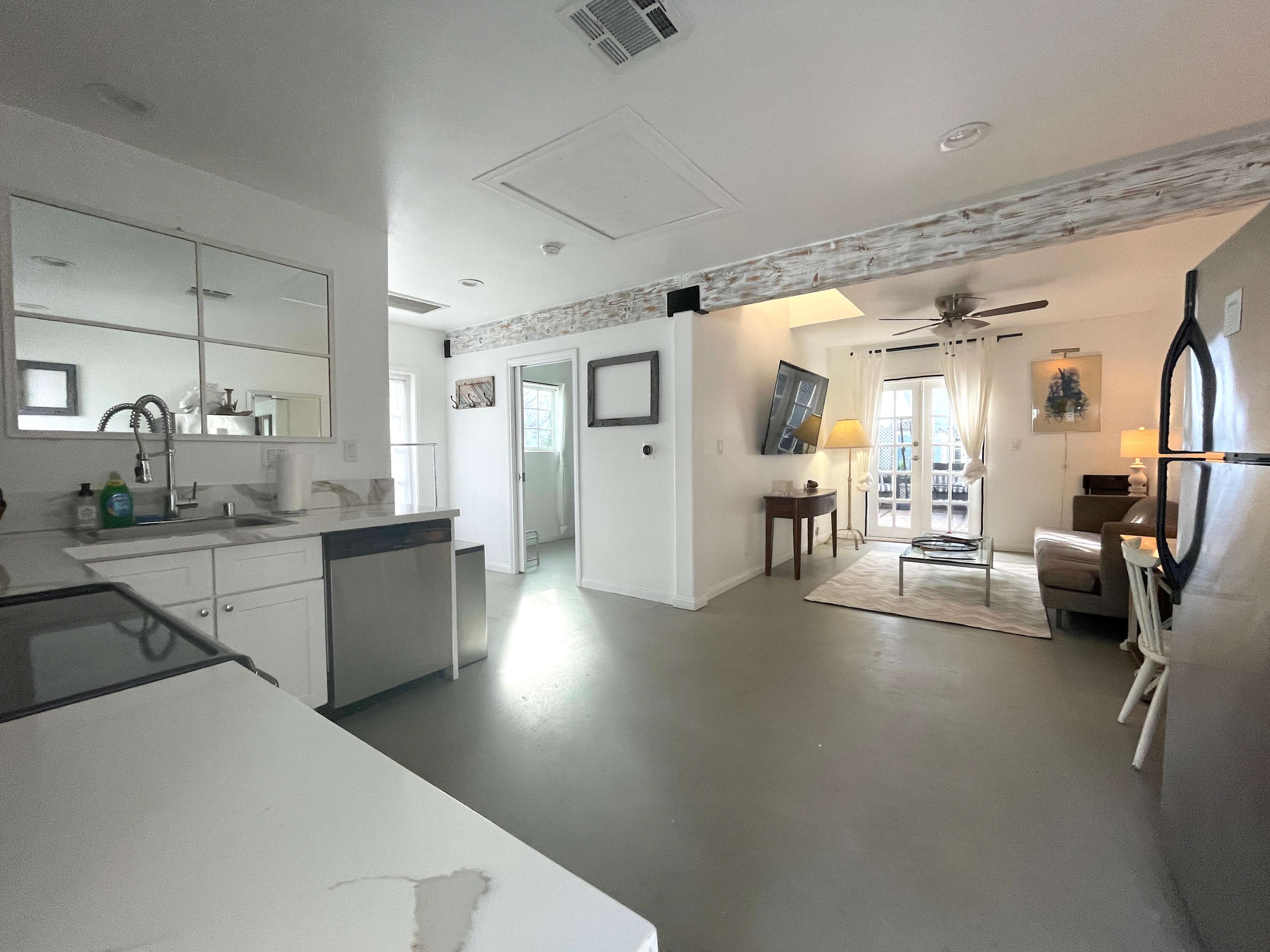 The image shows a modern, open-concept living space with a kitchen area on the left, a small dining table, and a living room area with a sofa and natural light streaming through a door.