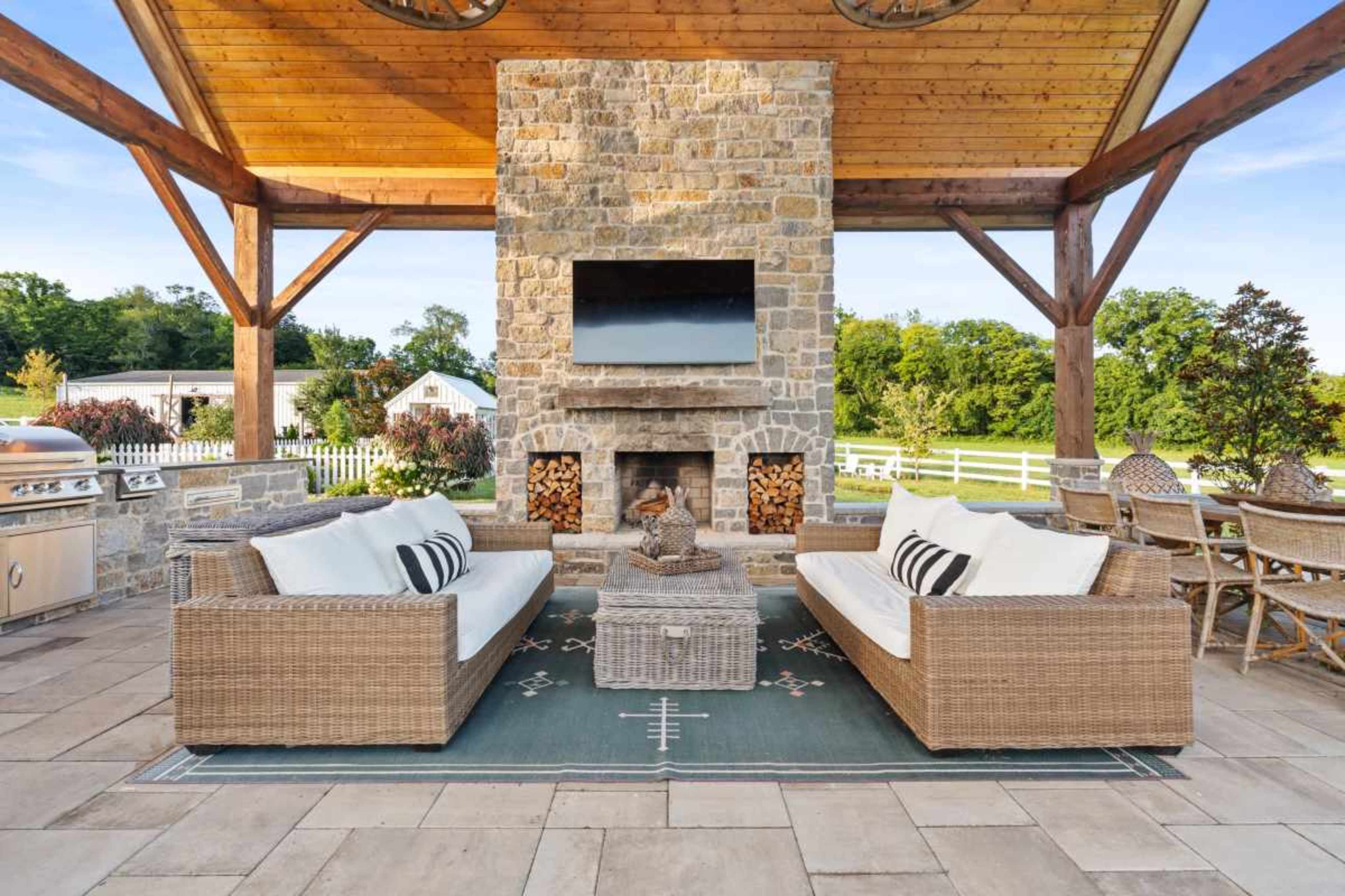 The image shows a spacious outdoor living area featuring comfortable wicker sofas, a stone fireplace with a mounted TV, and a view of greenery in the background.