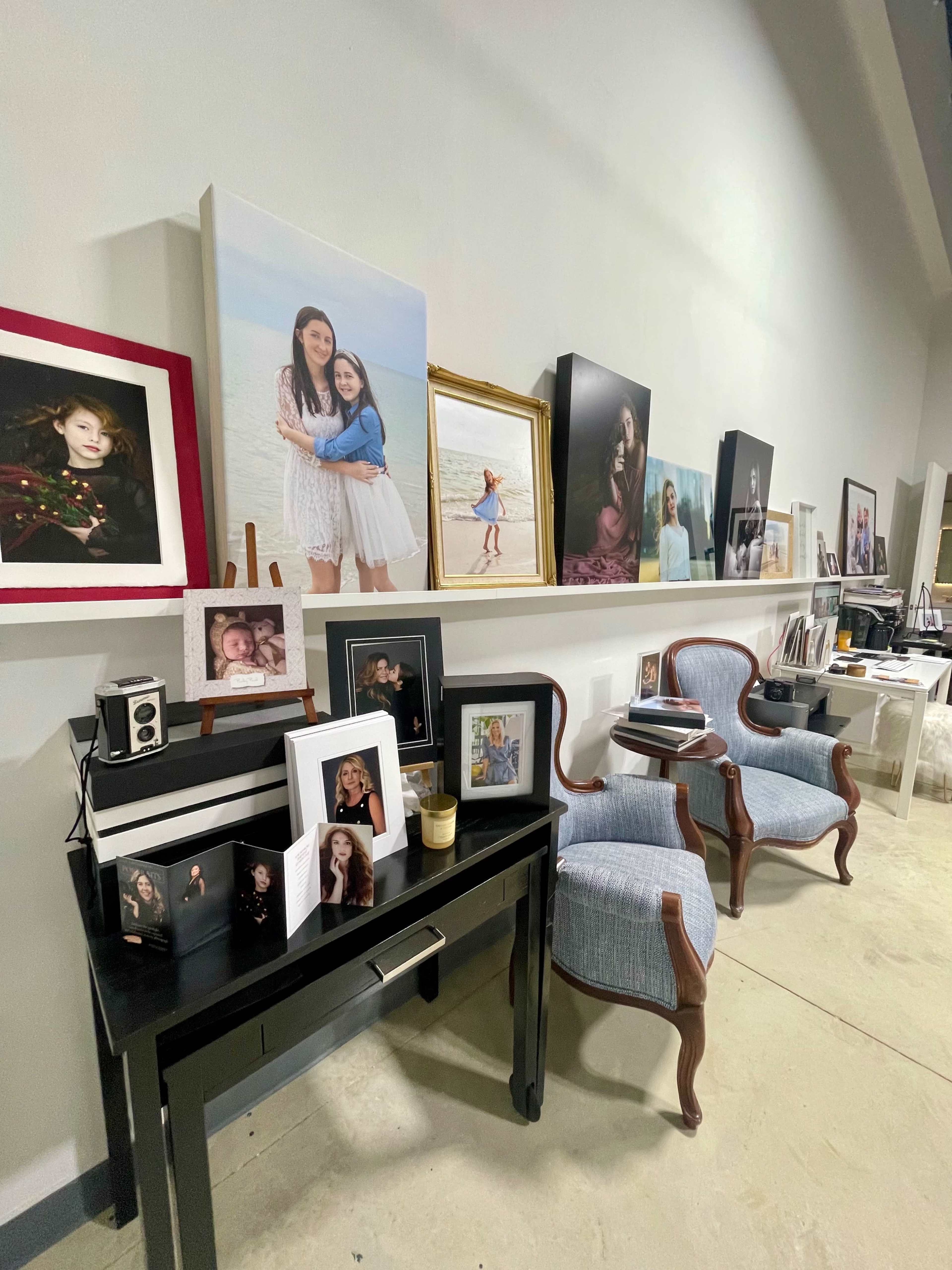 A studio space with a shelf displaying various framed photographs and two upholstered chairs beside a black console table.