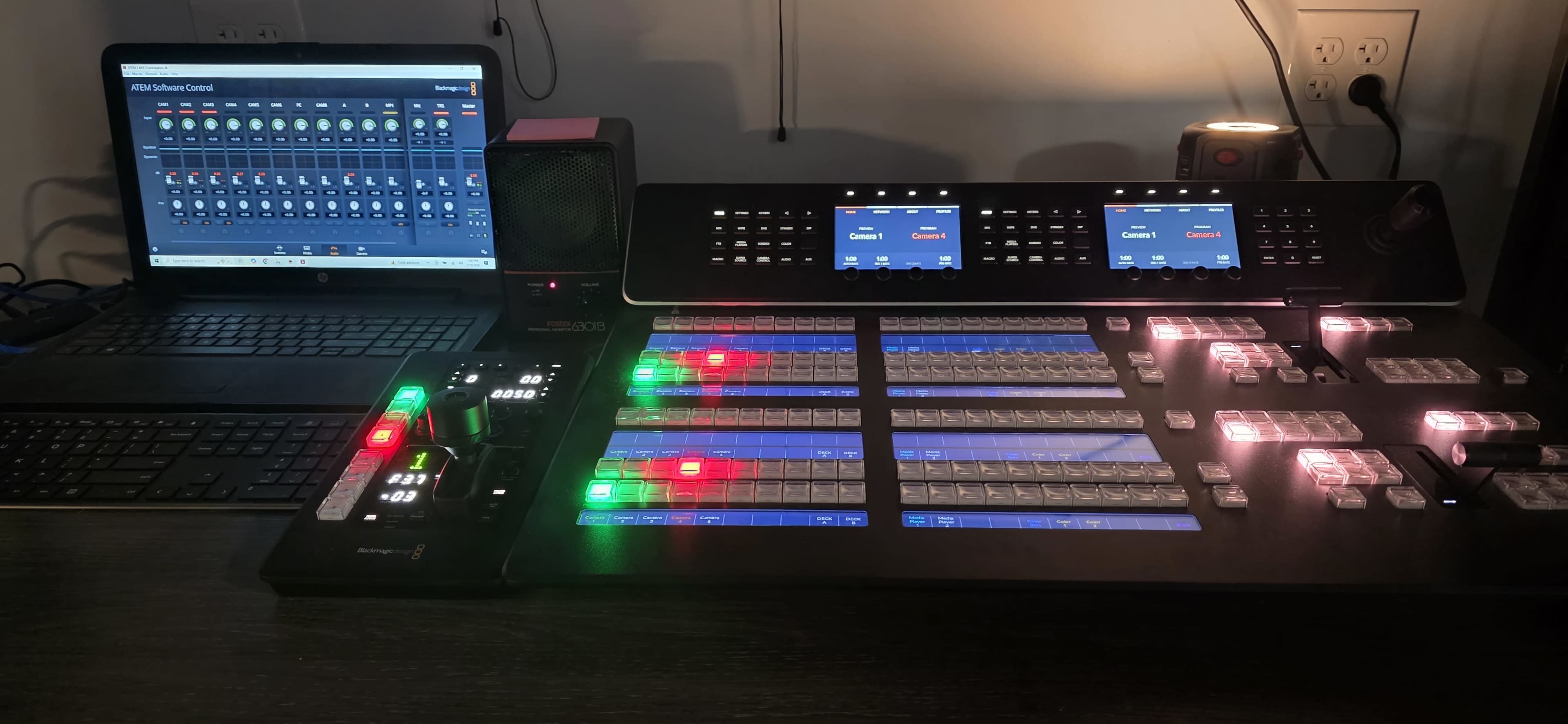 The image shows a video switcher console with illuminated buttons and a connected laptop displaying a control interface for video production.