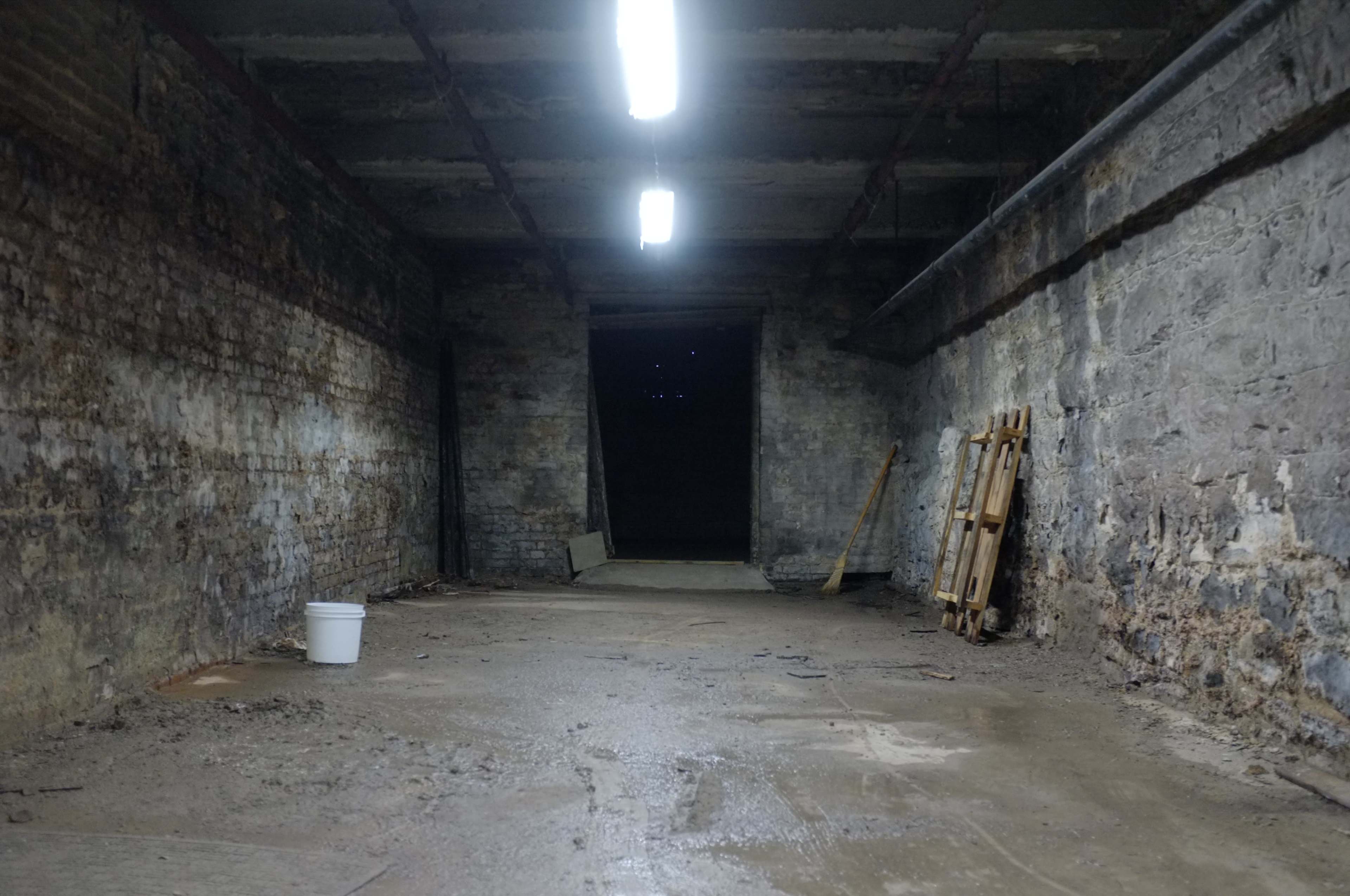 An empty, dimly lit, concrete room with exposed brick walls and a doorway leading to darkness.