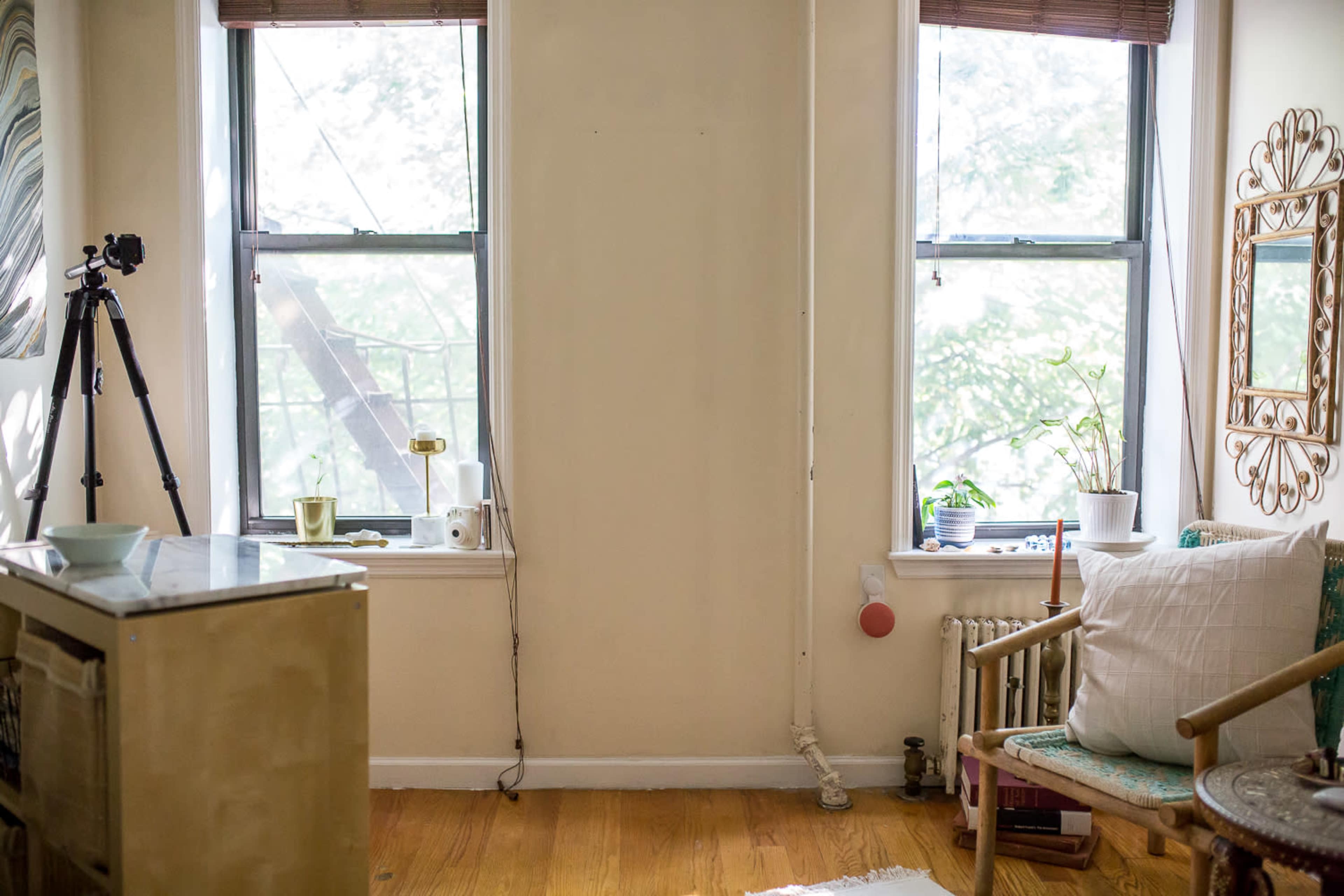 A well-lit corner of a room features two windows, a tripod, a small table with a few items, and plants on the sills.
