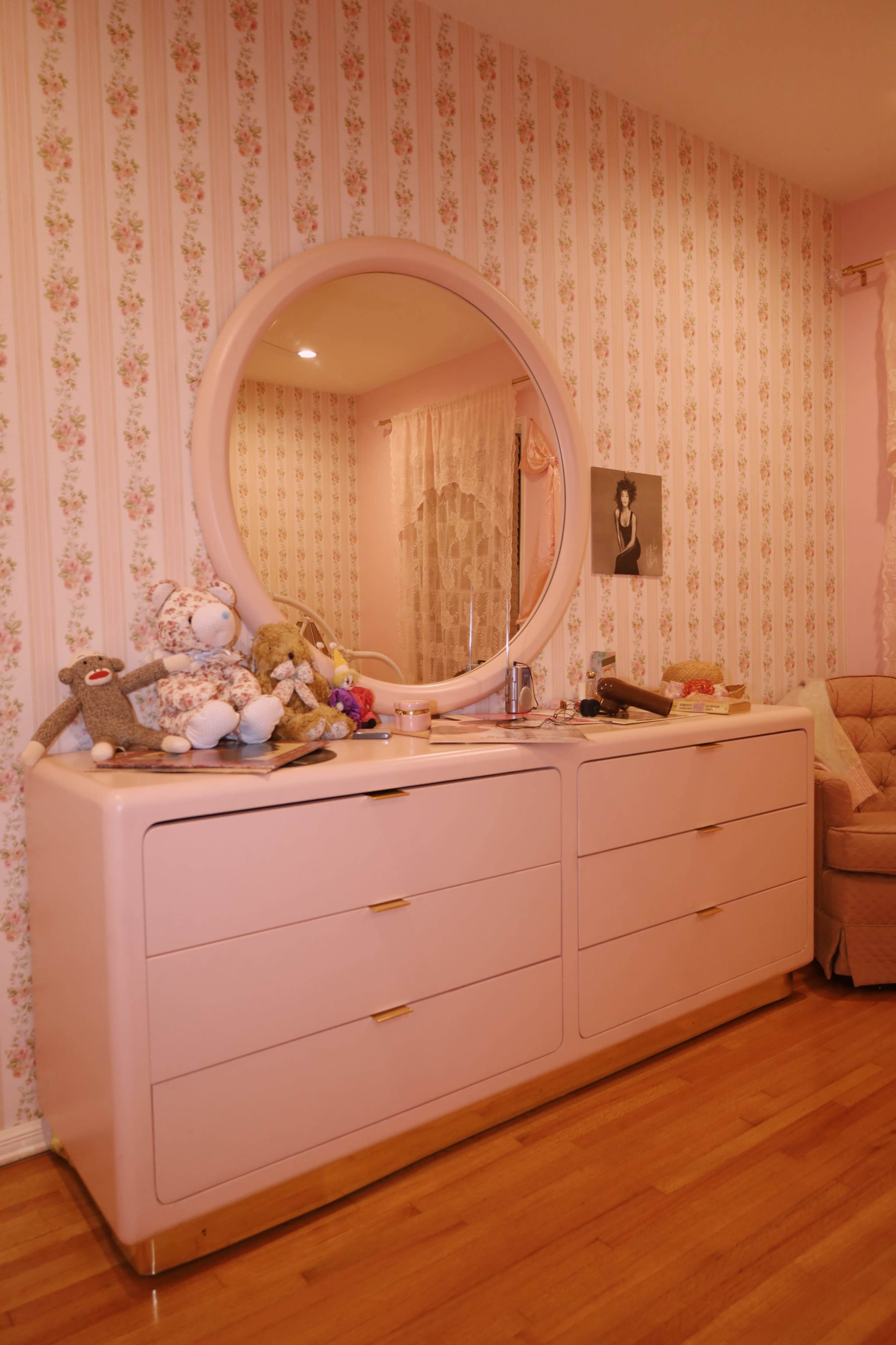A pink dresser with a round mirror sits against a floral-patterned wallpapered wall, cluttered with stuffed animals and various items.