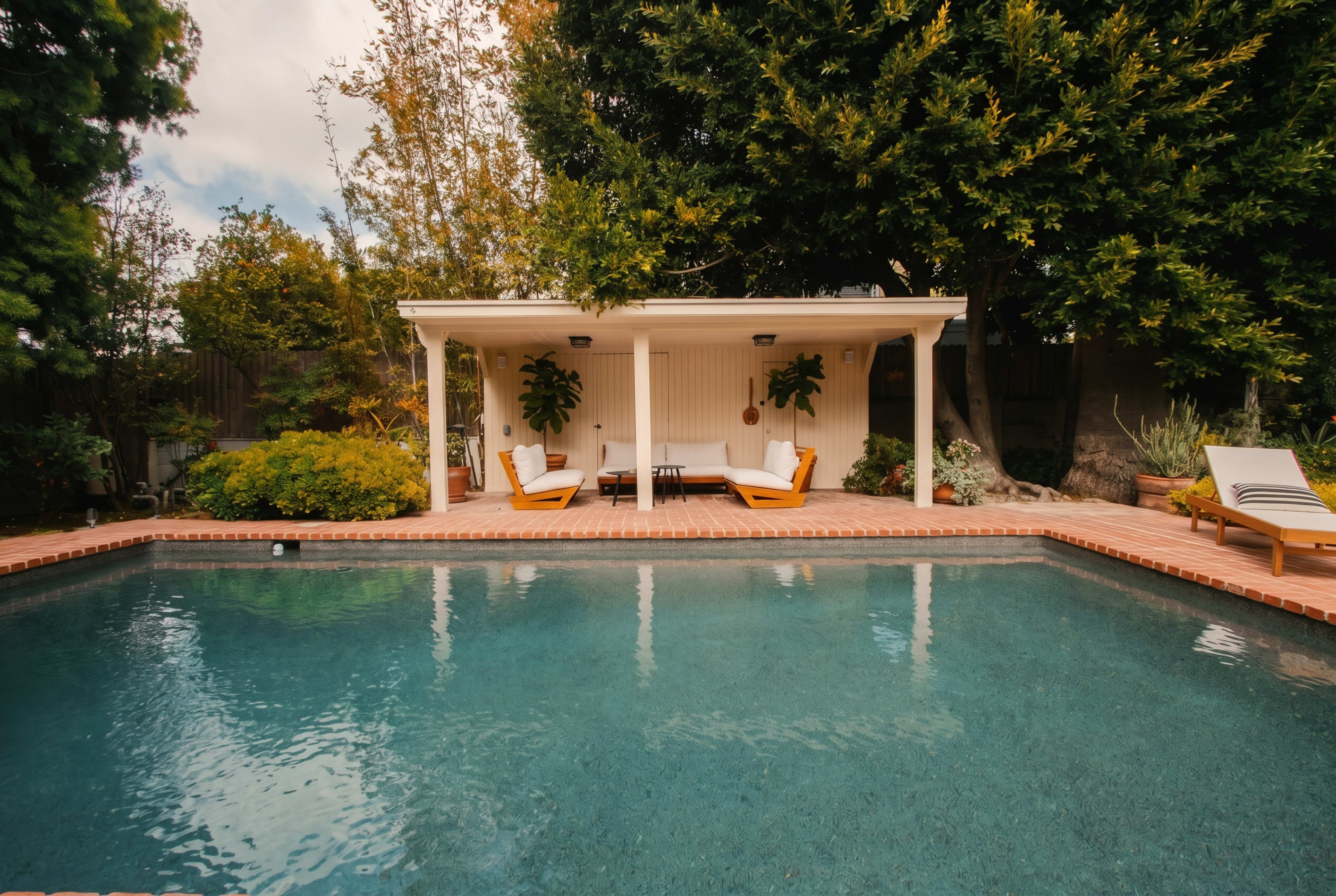 A swimming pool is situated in a backyard with lounge chairs and a covered seating area surrounded by greenery.