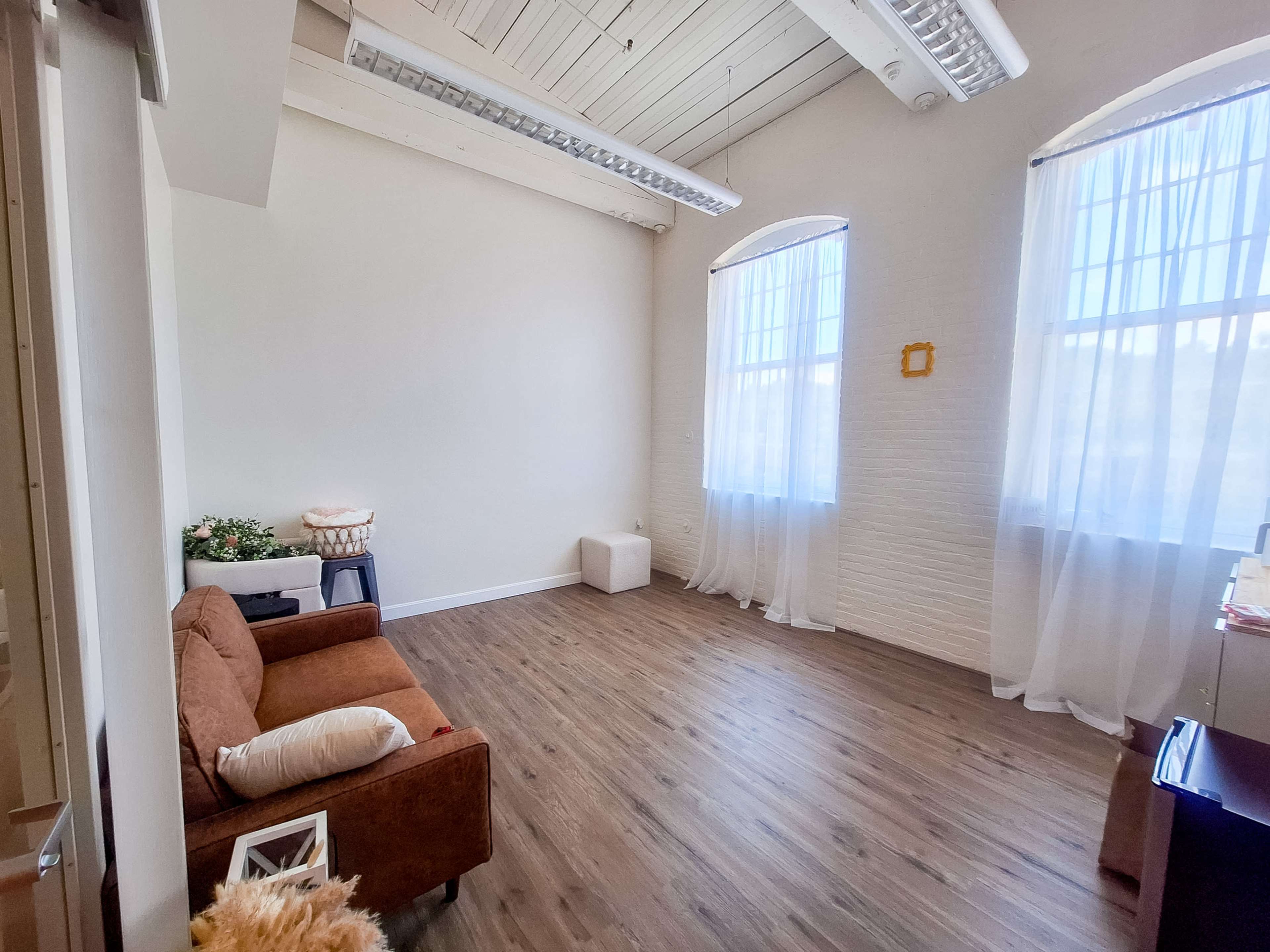 The image shows a spacious, well-lit room with two large windows, a brown sofa, and wooden flooring.