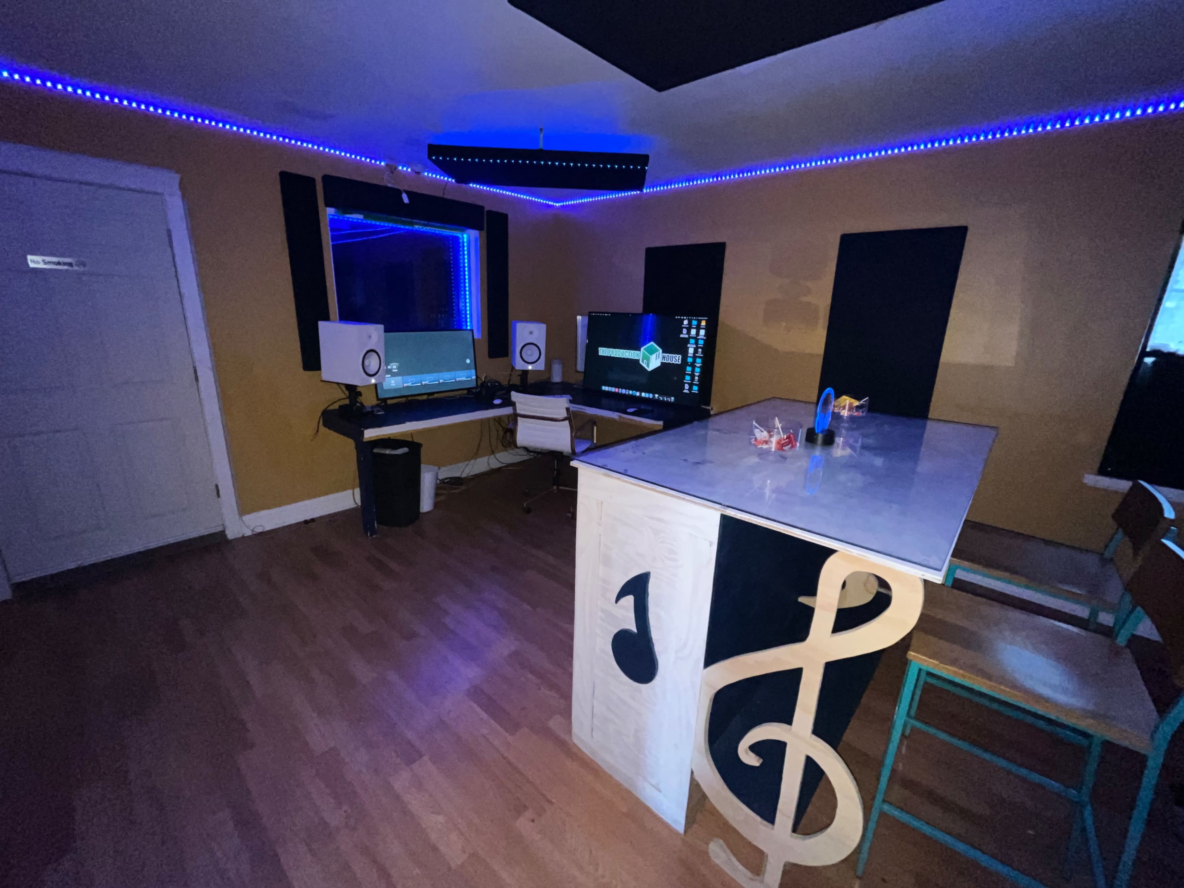 A small music studio featuring a desk with two monitors, studio speakers, and colorful LED strip lighting along the walls.