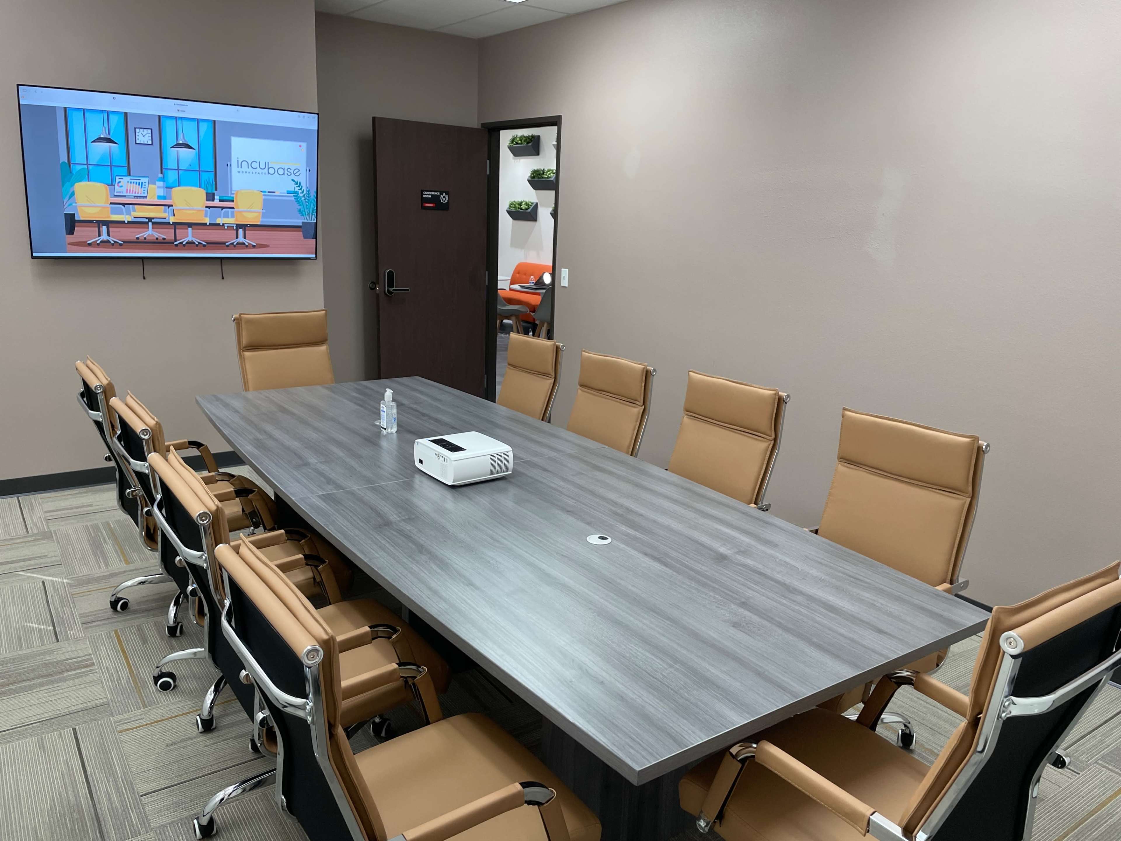 A conference room features a large gray table surrounded by multiple brown chairs, with a projector on the table and a screen displaying a colorful presentation on the wall.
