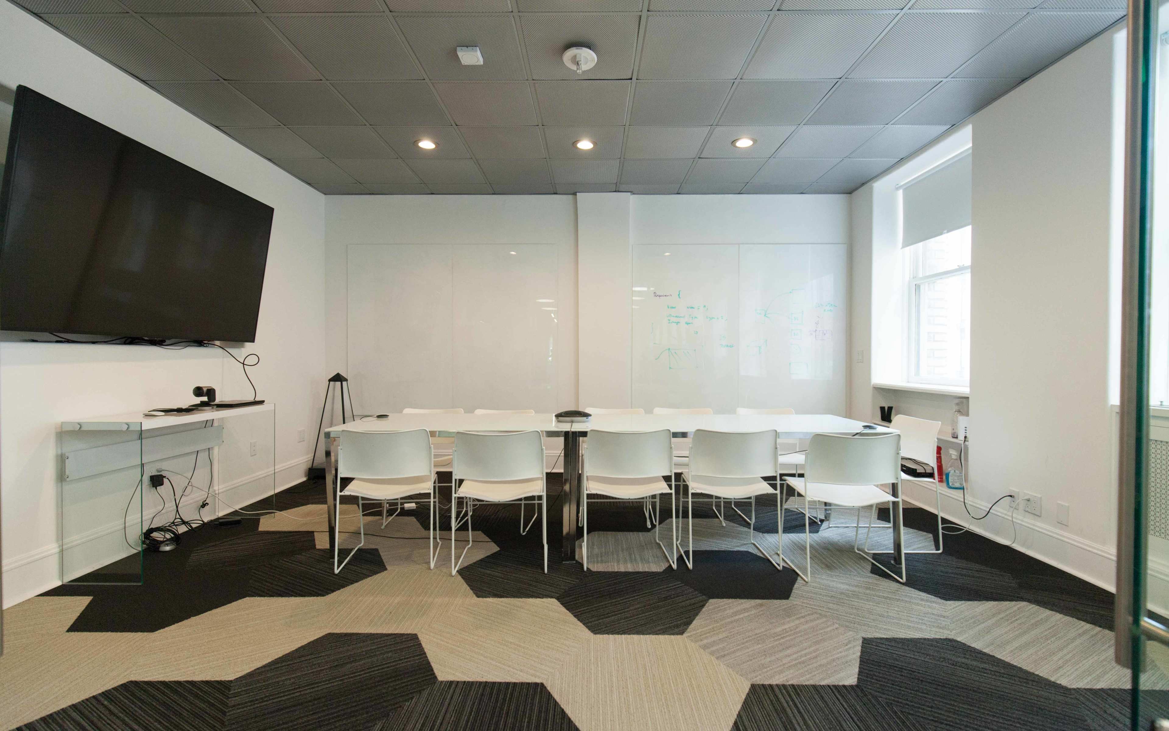 A modern conference room features a long table surrounded by white chairs and a large TV mounted on the wall.
