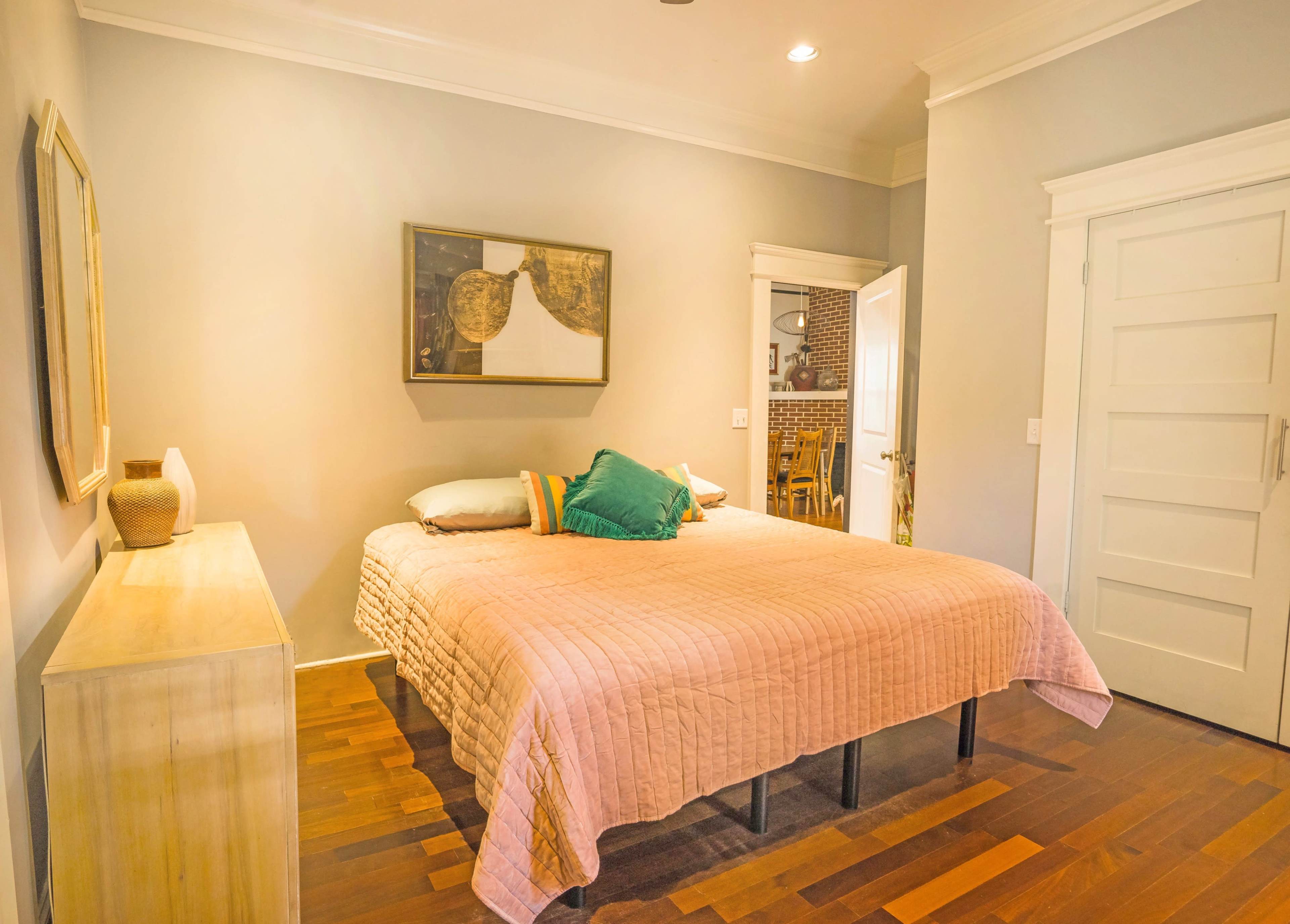 A neatly arranged bedroom with a large bed covered in a pink quilt, a wooden dresser, and a doorway leading to another room.