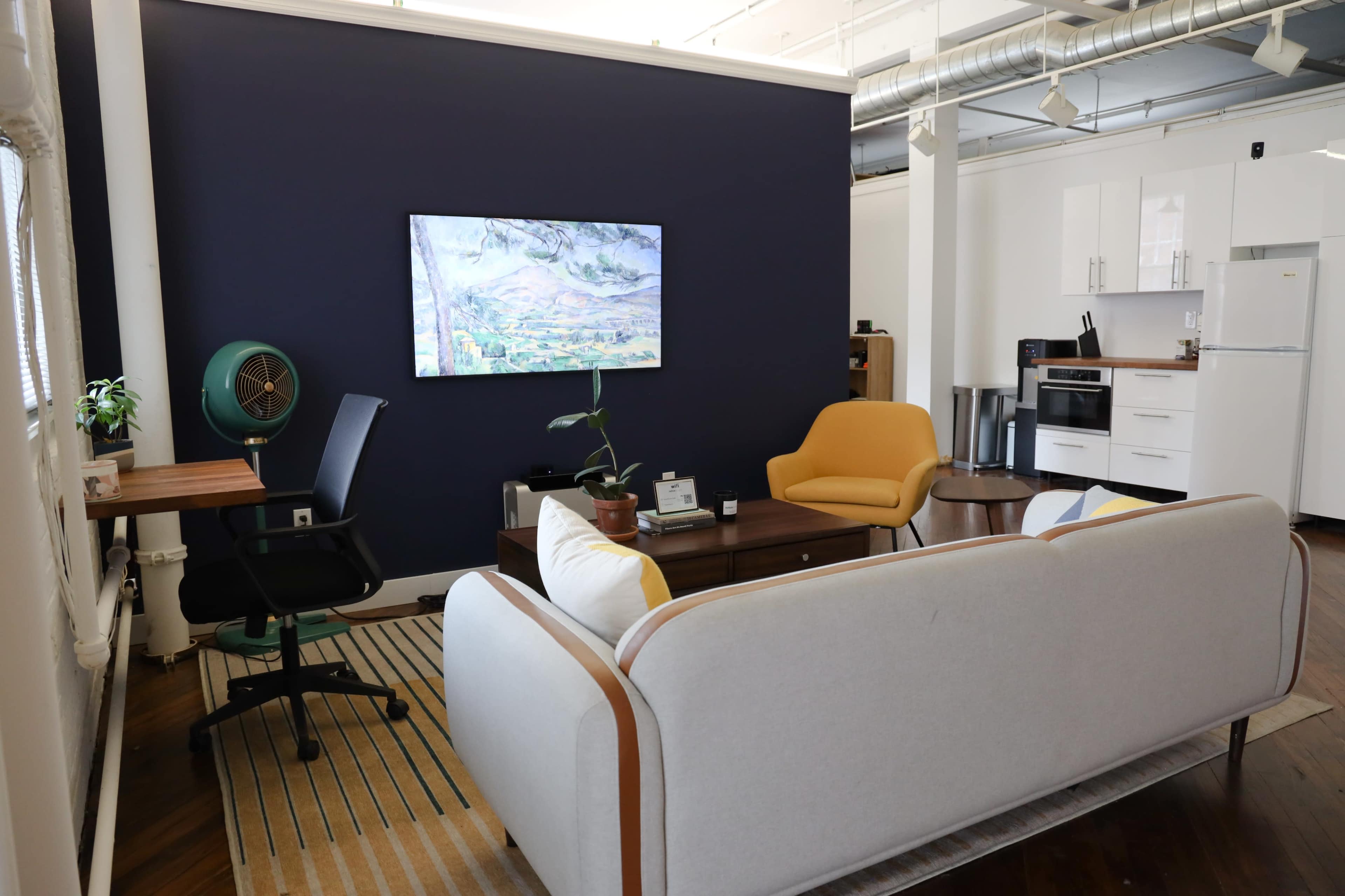 A modern living space featuring a light-colored sofa, a yellow chair, a television on the wall, and a kitchen area with white cabinets.