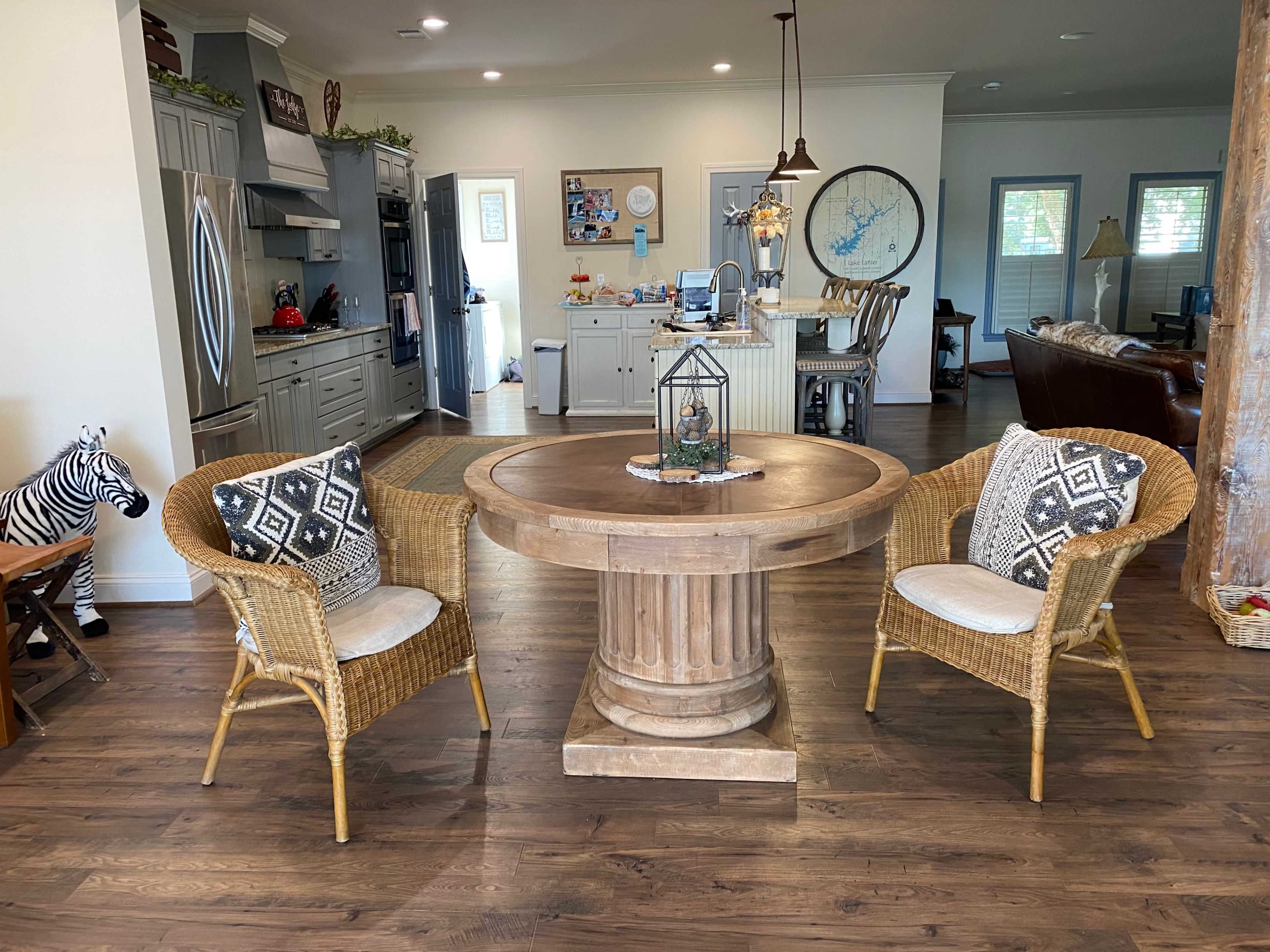 A round wooden table with a pedestal base is centered between two wicker chairs adorned with patterned cushions in a well-lit living space.