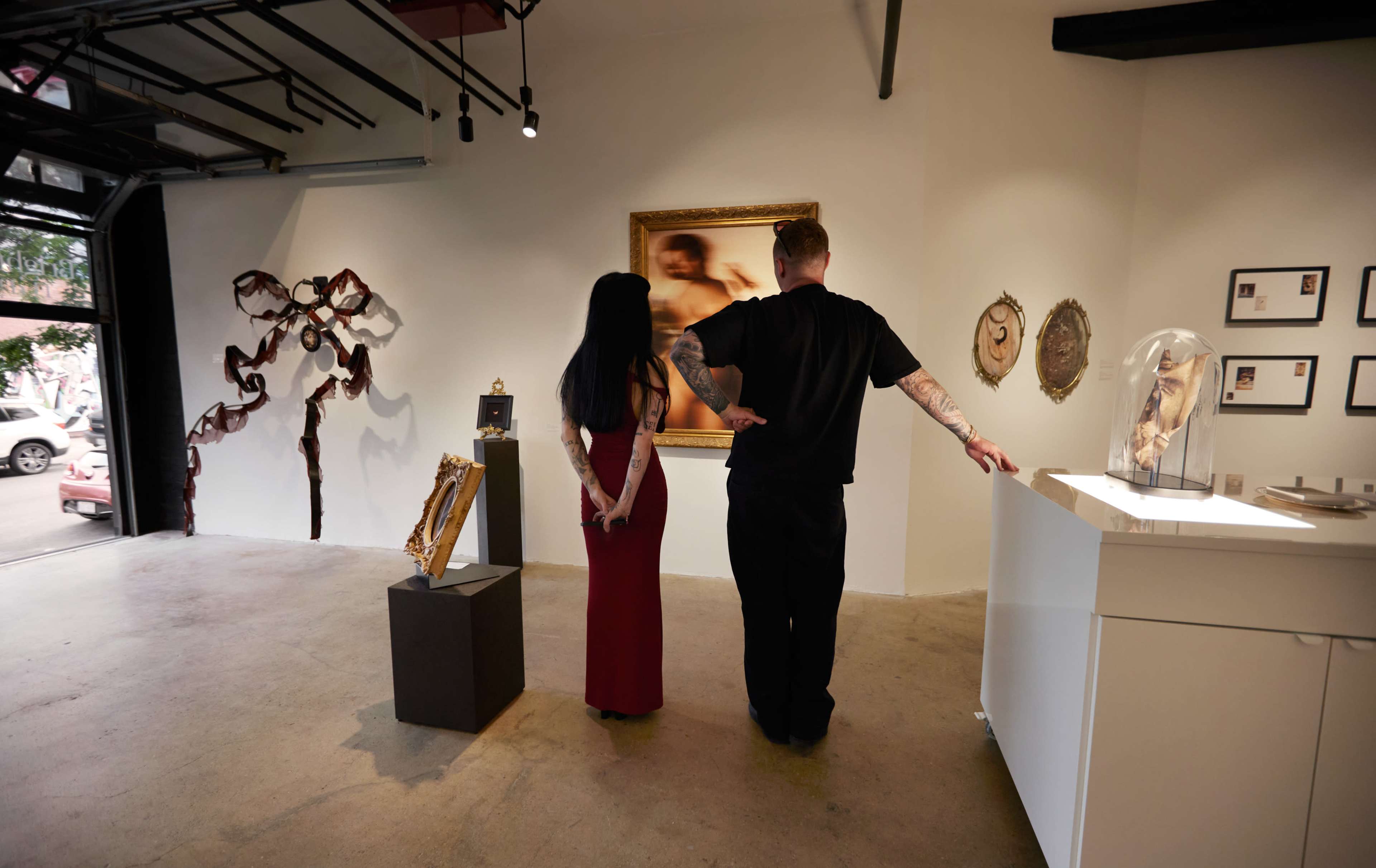 A woman in a red dress and a man in black stand together in an art gallery, examining framed artwork on the wall.