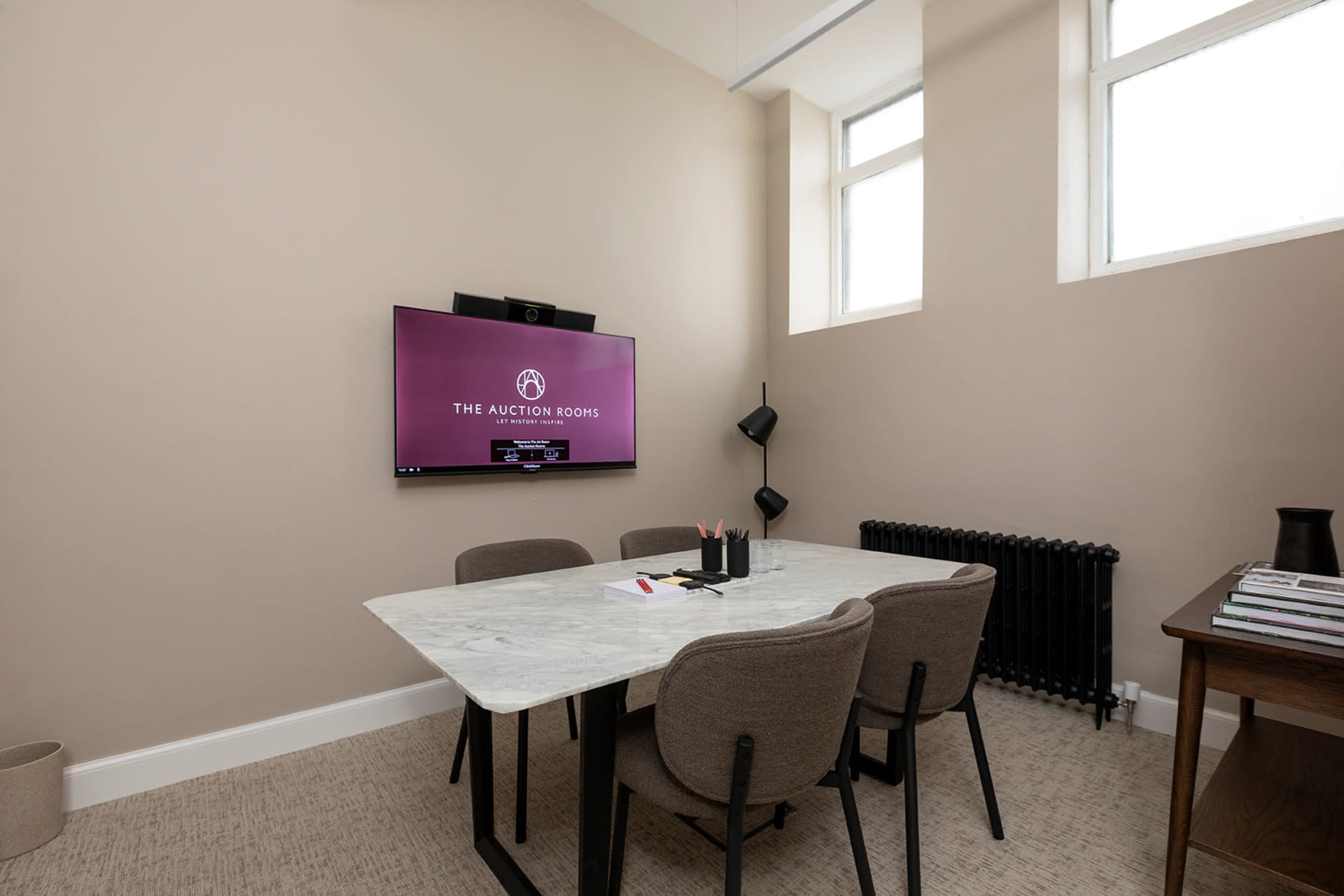 A small conference room features a marble table surrounded by four chairs, with a wall-mounted screen displaying "THE AUCTION ROOMS," and a radiator visible in the background.