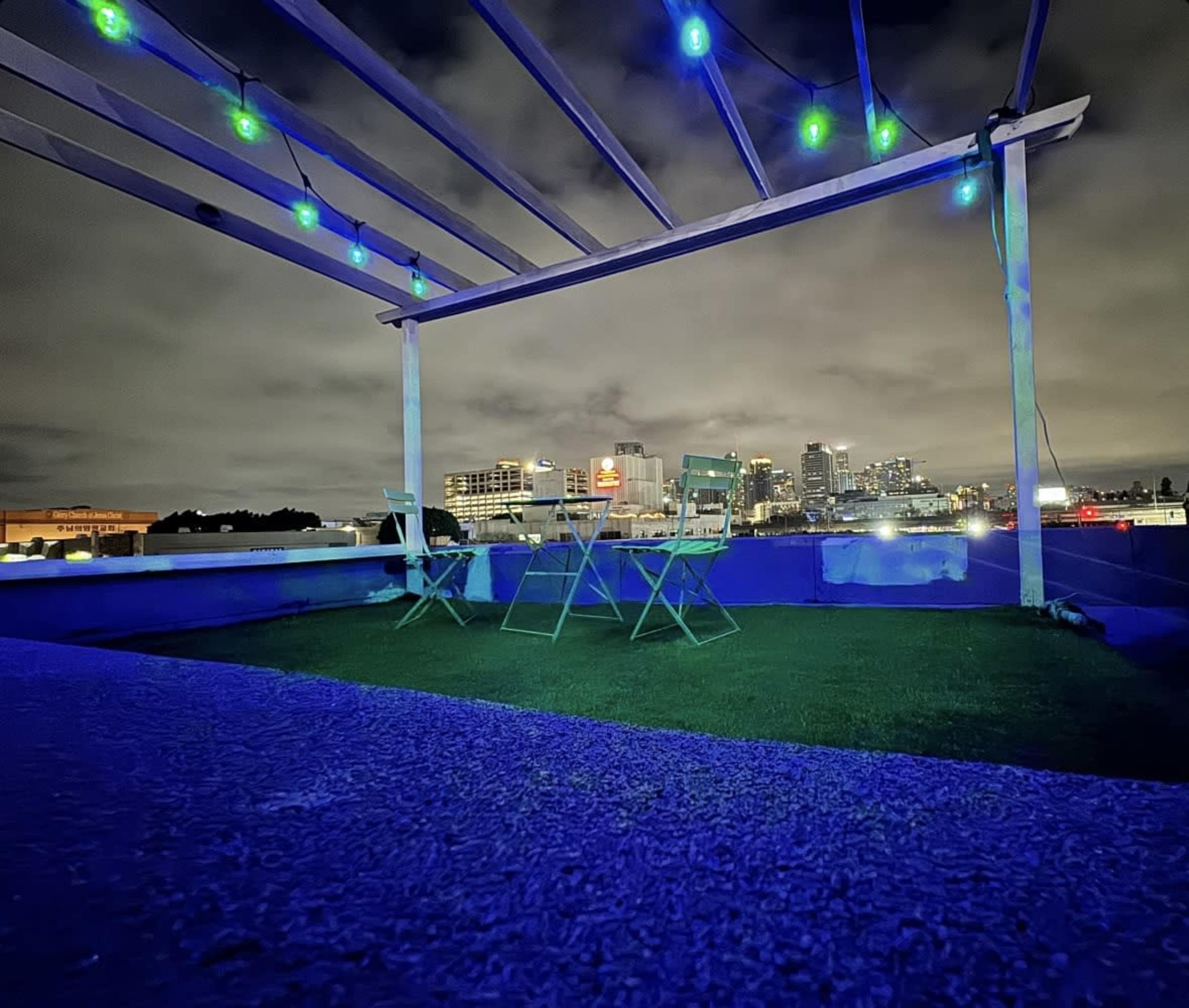 The image shows a rooftop space with green chairs under a lighted pergola, overlooking a city skyline at night.