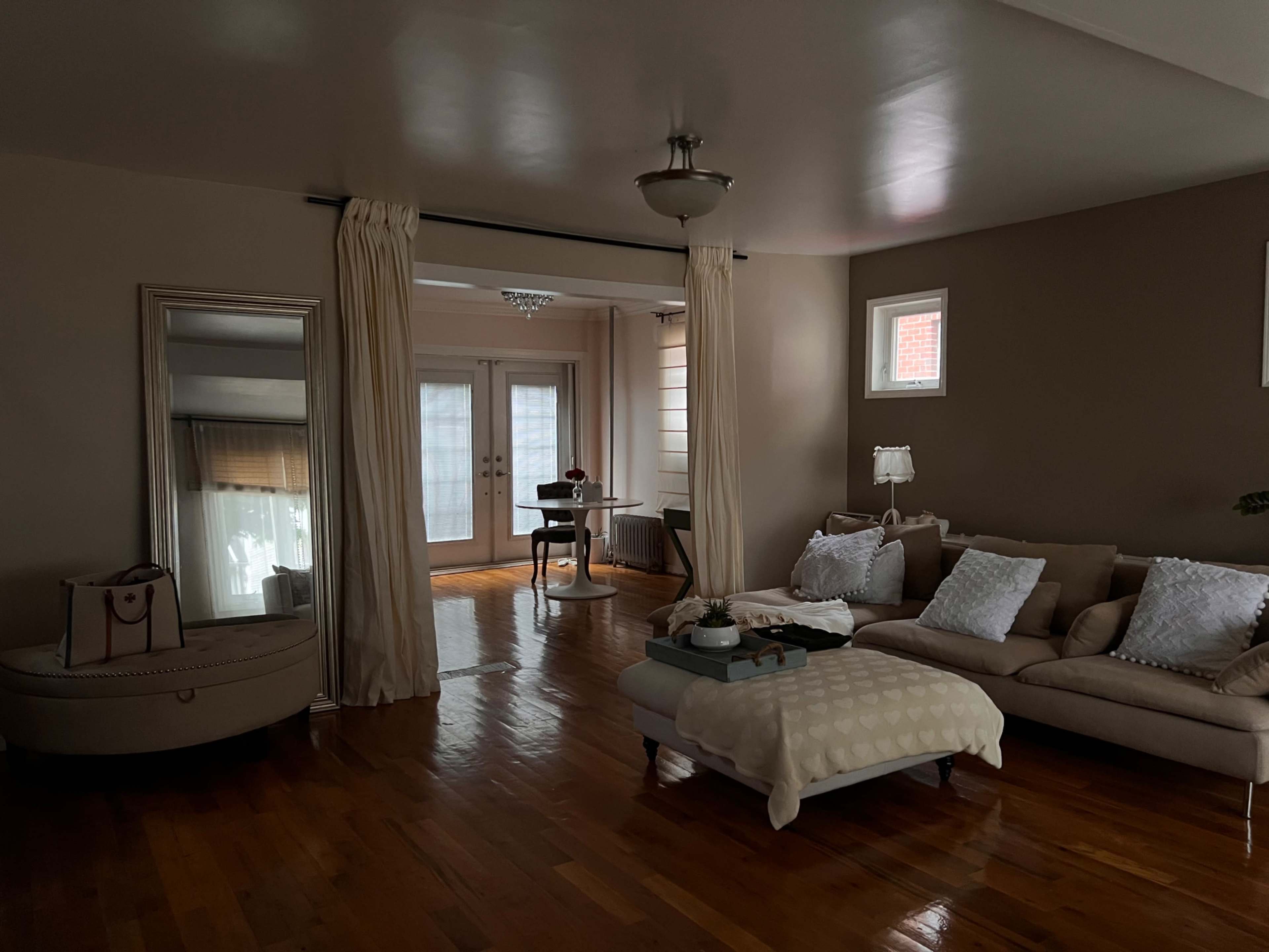 A spacious living room features a sectional sofa, a round dining table, and a large mirror, with sunlight streaming through the windows.
