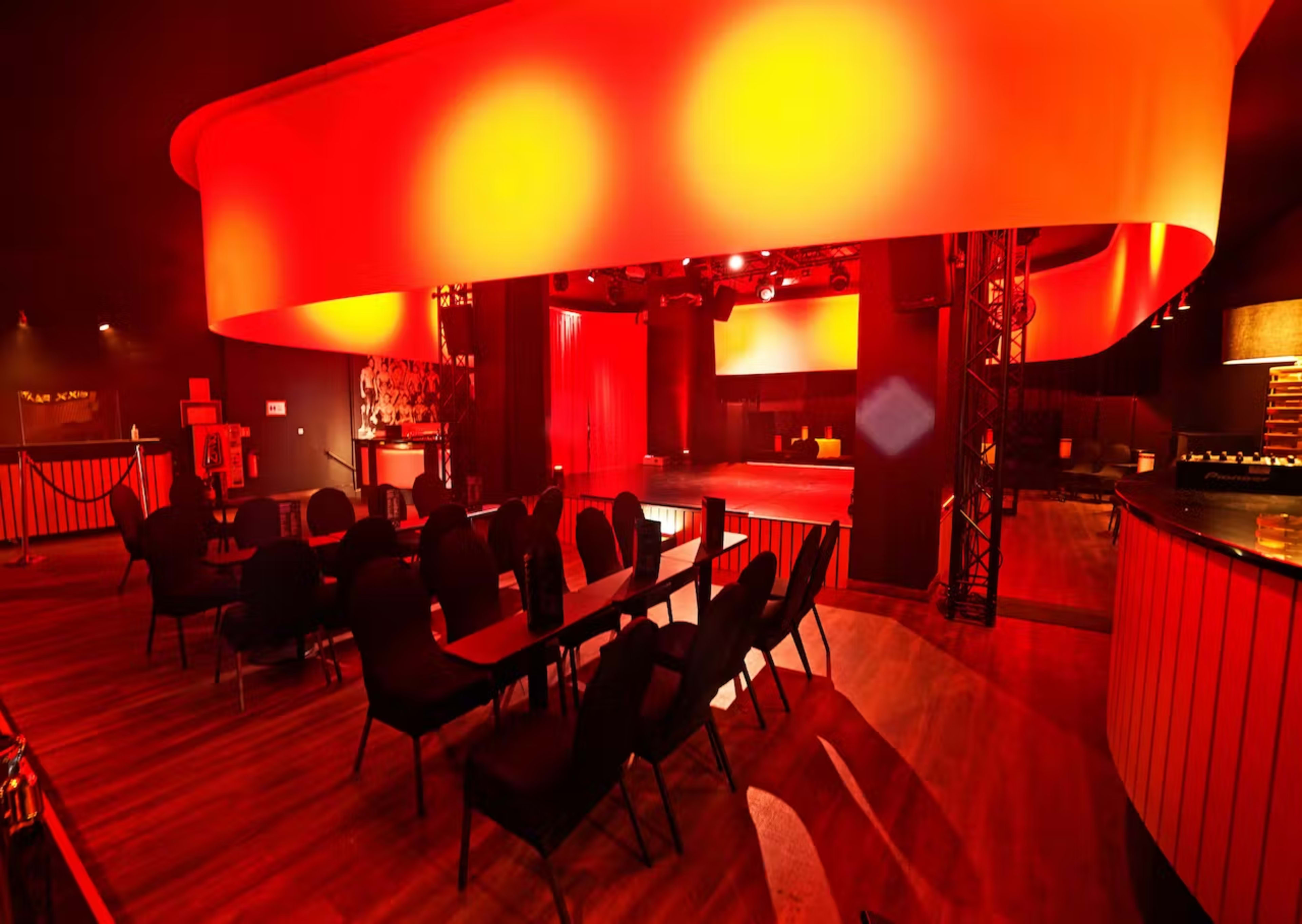 The image shows a dimly lit performance venue featuring a circular overhead light, rows of black chairs, and a stage area with red curtains.