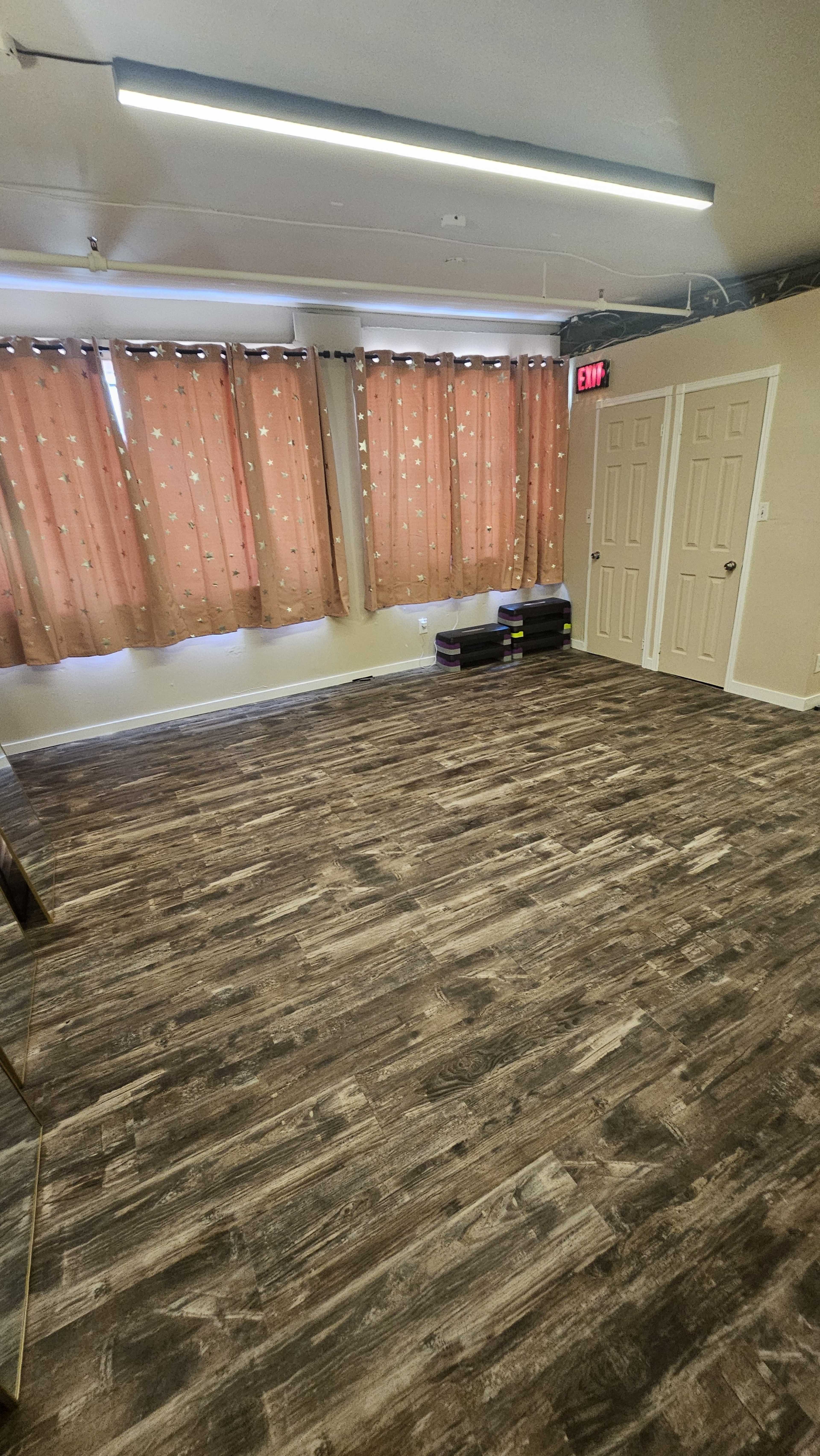 The image shows a room with wooden flooring, beige walls, and large windows covered by orange curtains, along with two closed doors on one side.