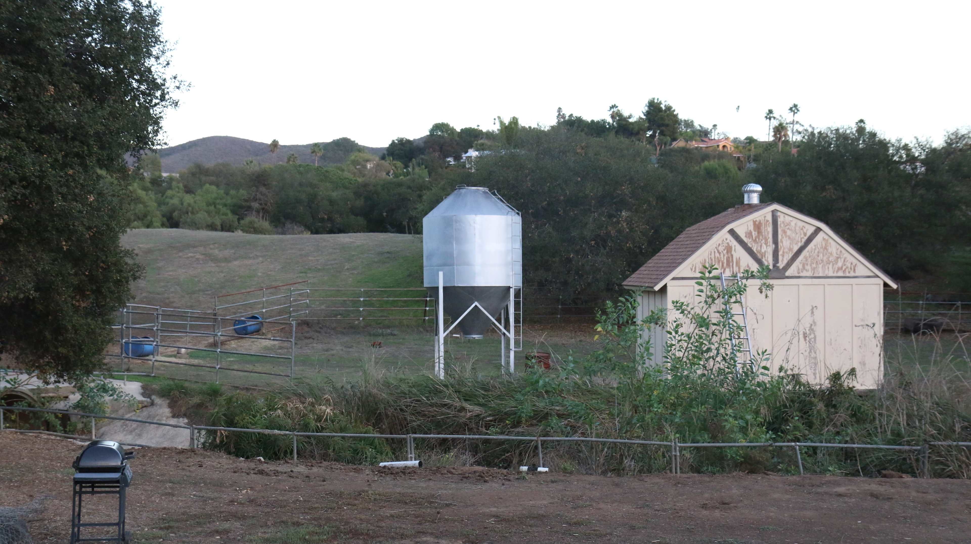 The image shows a rural landscape featuring a metal grain silo, a small shed, and a fenced area near a creek surrounded by greenery.