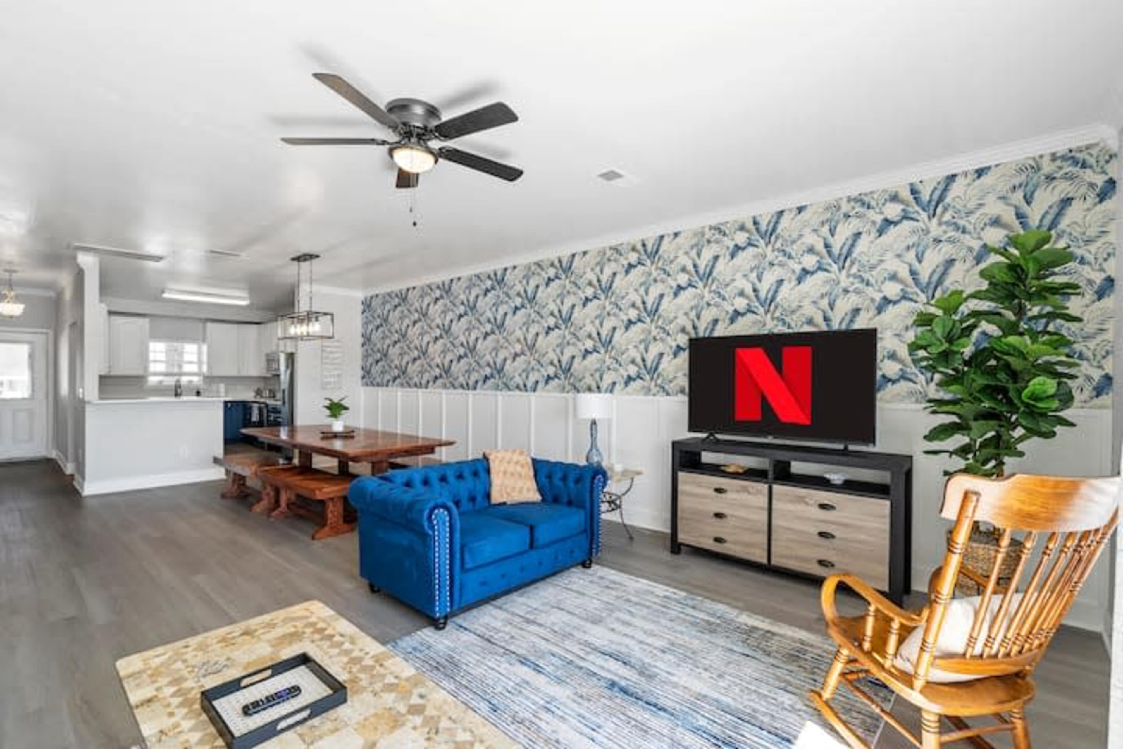 The image features a living room with a blue sofa, a large TV displaying the Netflix logo, a wooden dining table, and a patterned wallpaper.