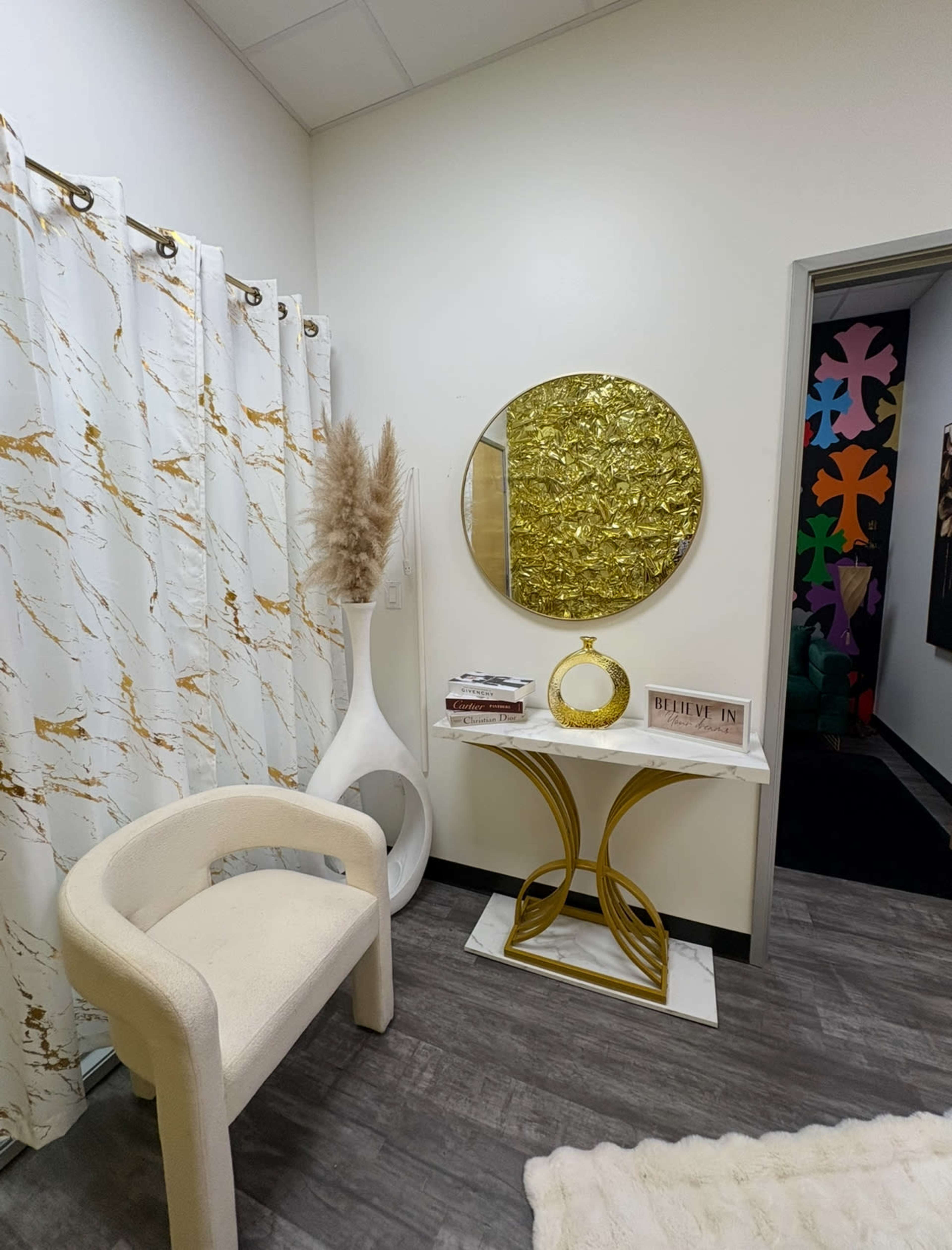 The image shows a modern interior corner featuring a white chair, a round gold mirror, a decorative table with gold accents, and a large vase with dried pampas grass, set against a backdrop of marble-patterned curtains.