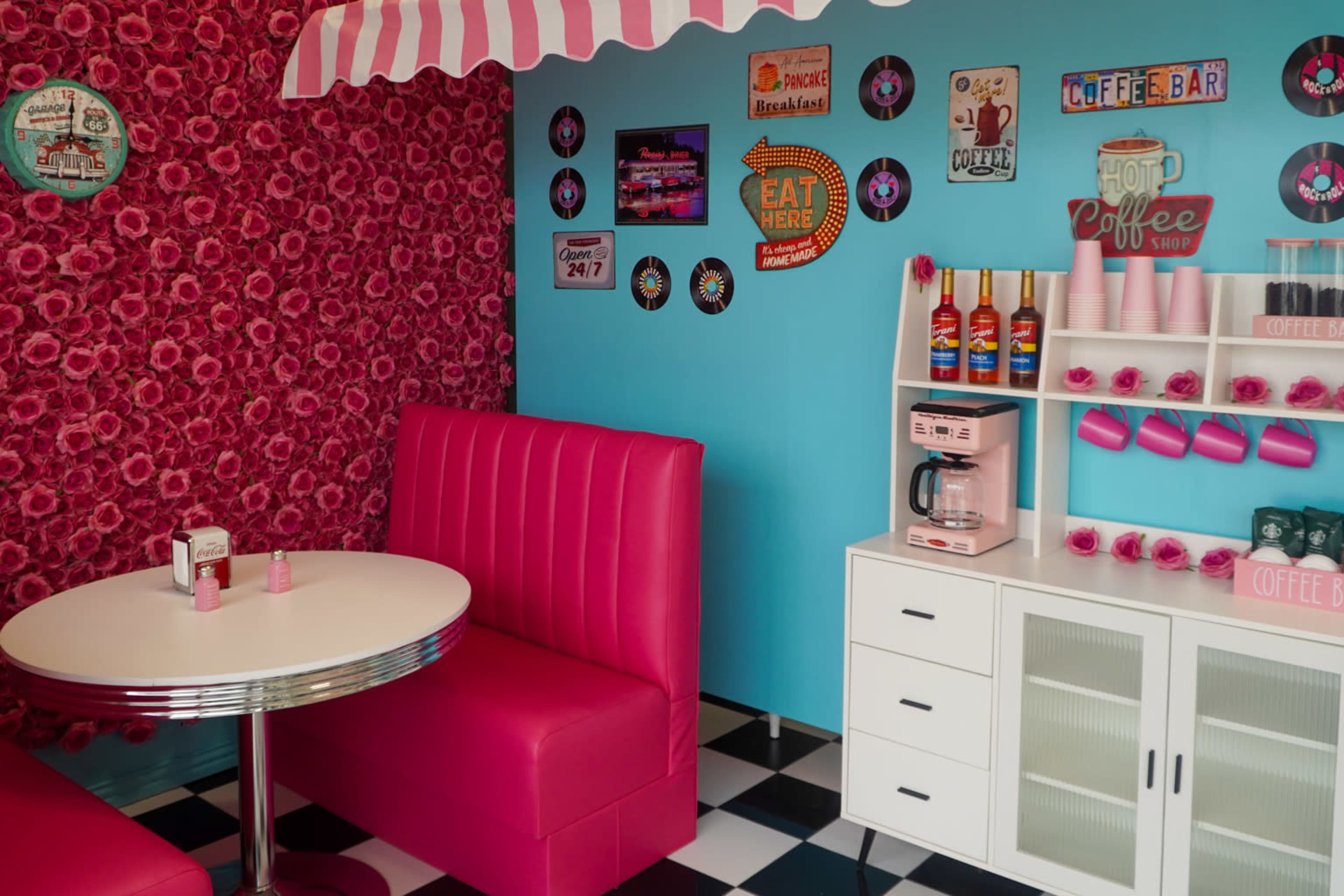 The image shows a retro-style diner corner with pink seating, vintage decor, and a coffee station featuring colorful syrup bottles.