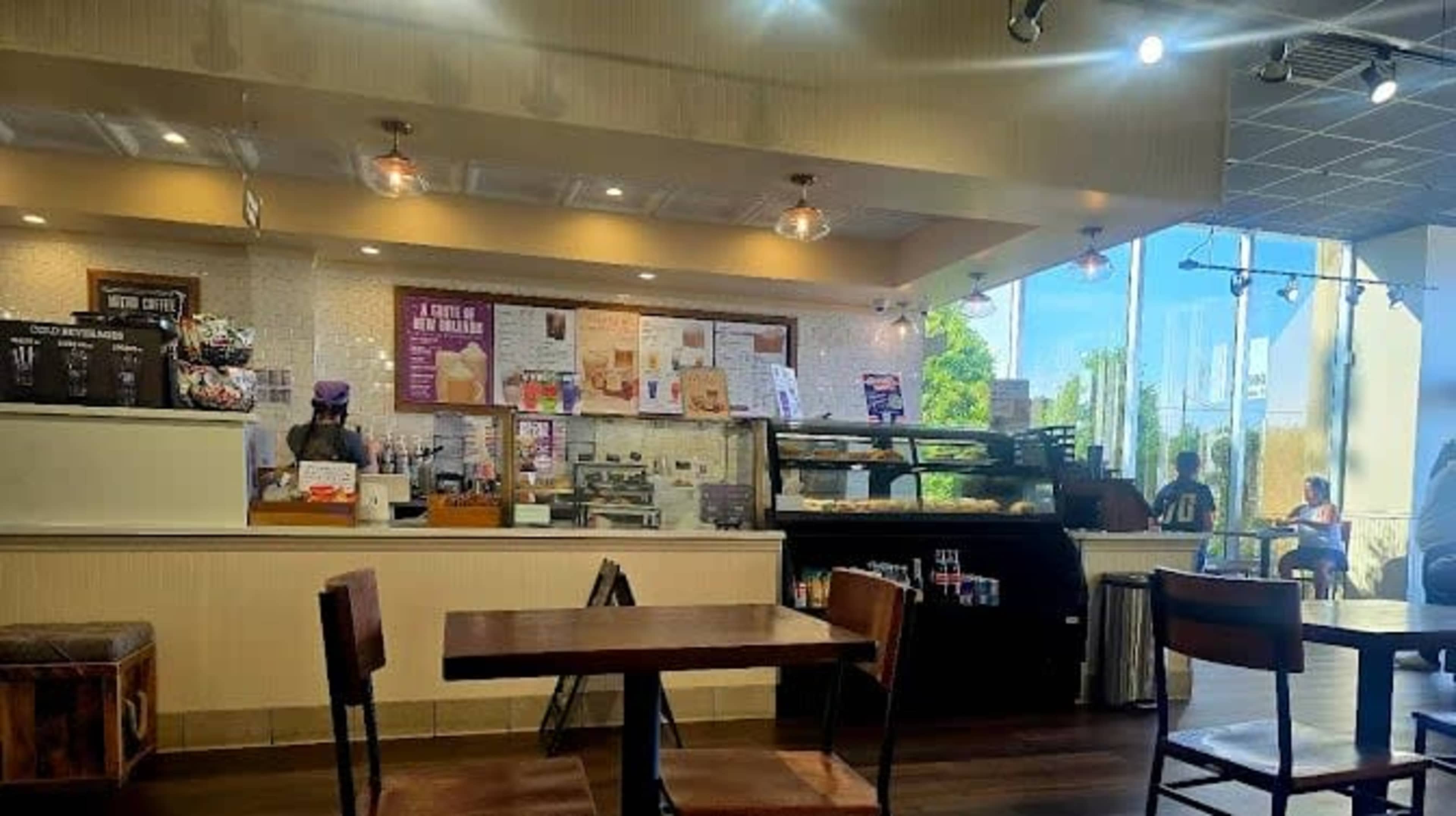 A coffee shop interior features a service counter with various beverages and snacks, surrounded by seating areas and large windows letting in natural light.