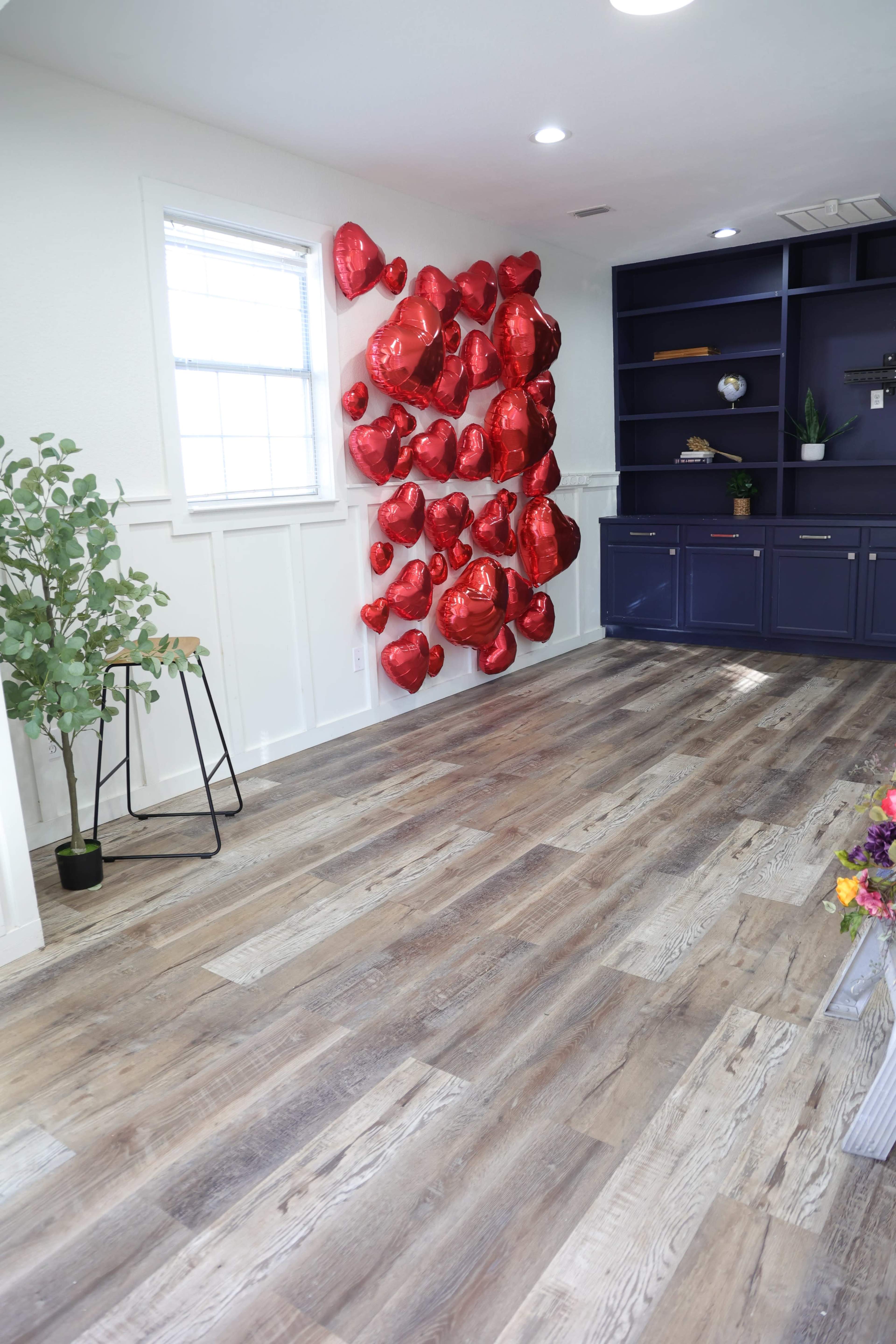 The image shows a room with wooden flooring and a wall adorned with red heart-shaped balloons, alongside a small plant and a stool.