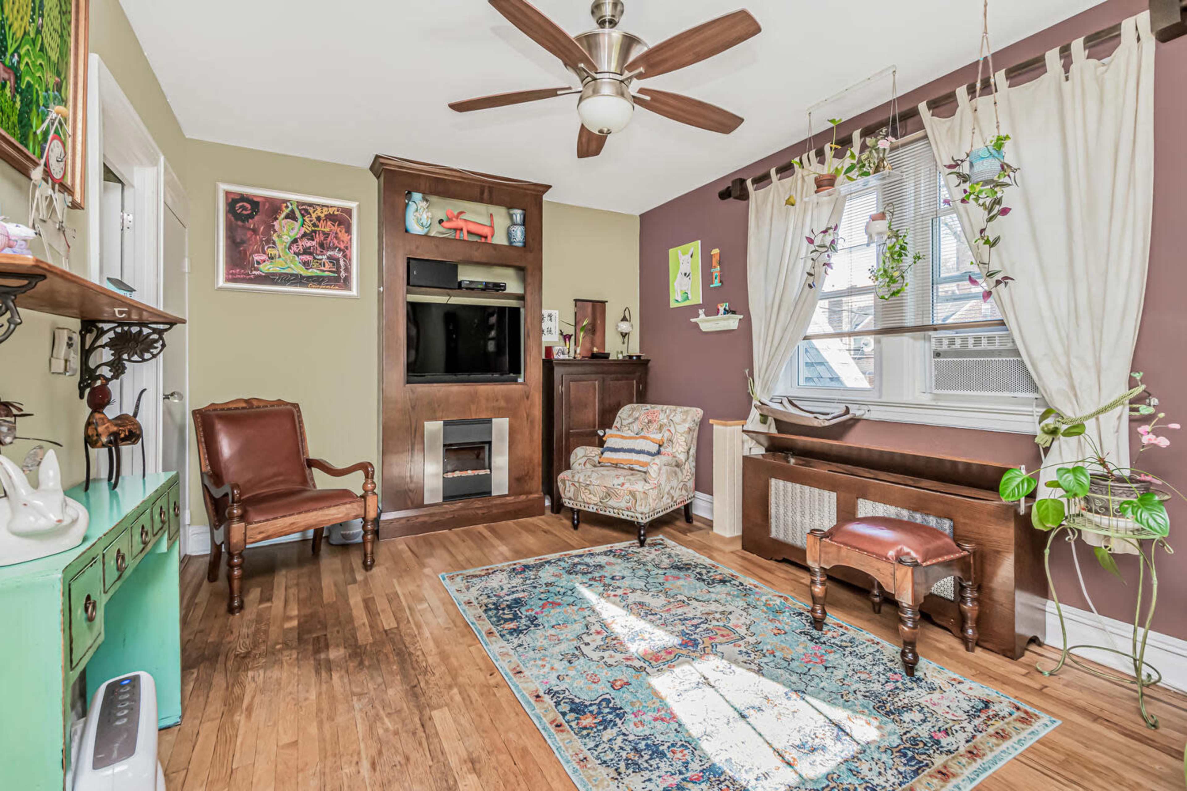 The living room features a hardwood floor, a mounted television unit, a wooden console, a small rug, and decorative plants hanging by the window.