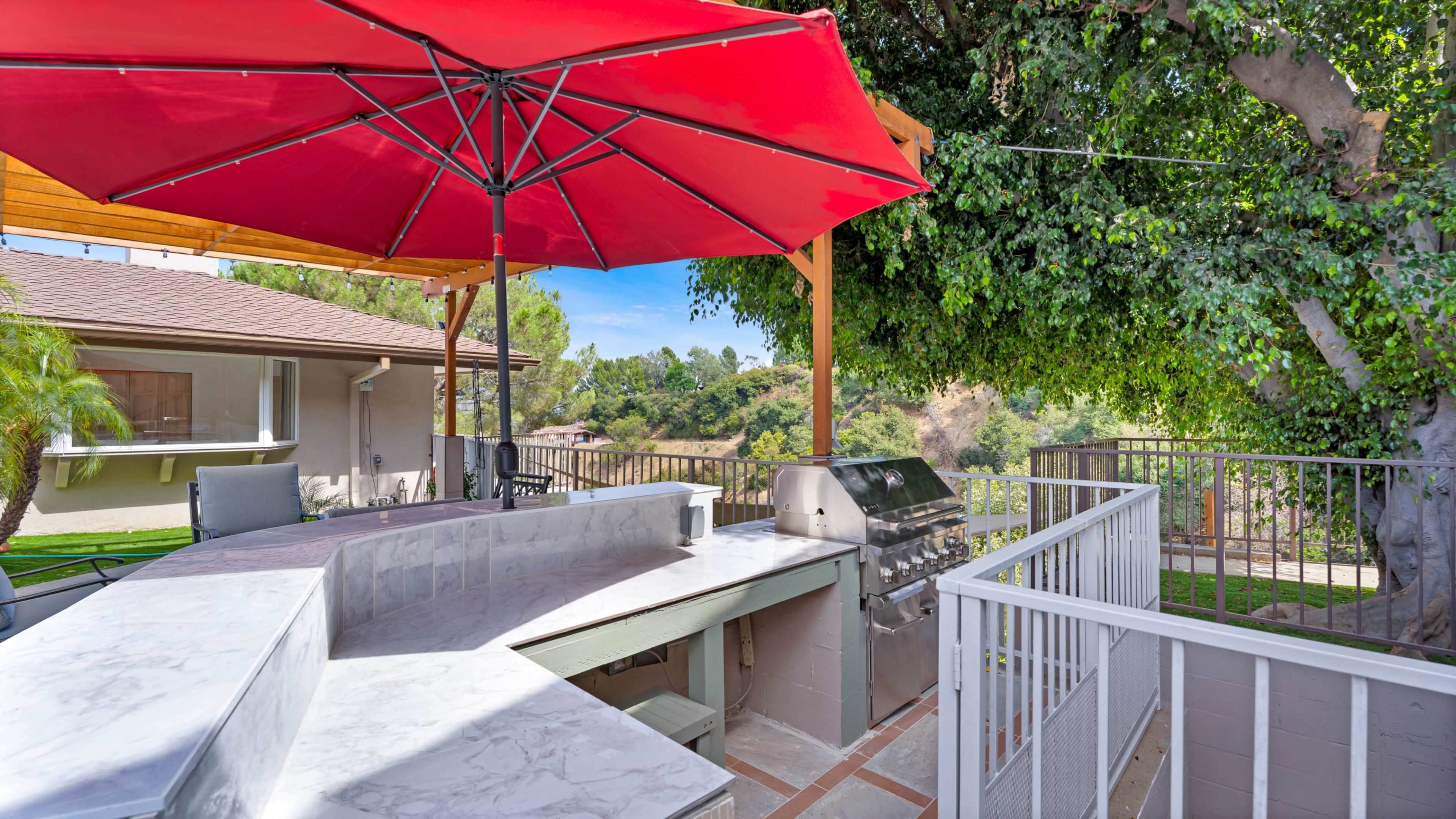 The image shows a backyard barbecue area with a marble countertop, a grill, and a large red umbrella providing shade.