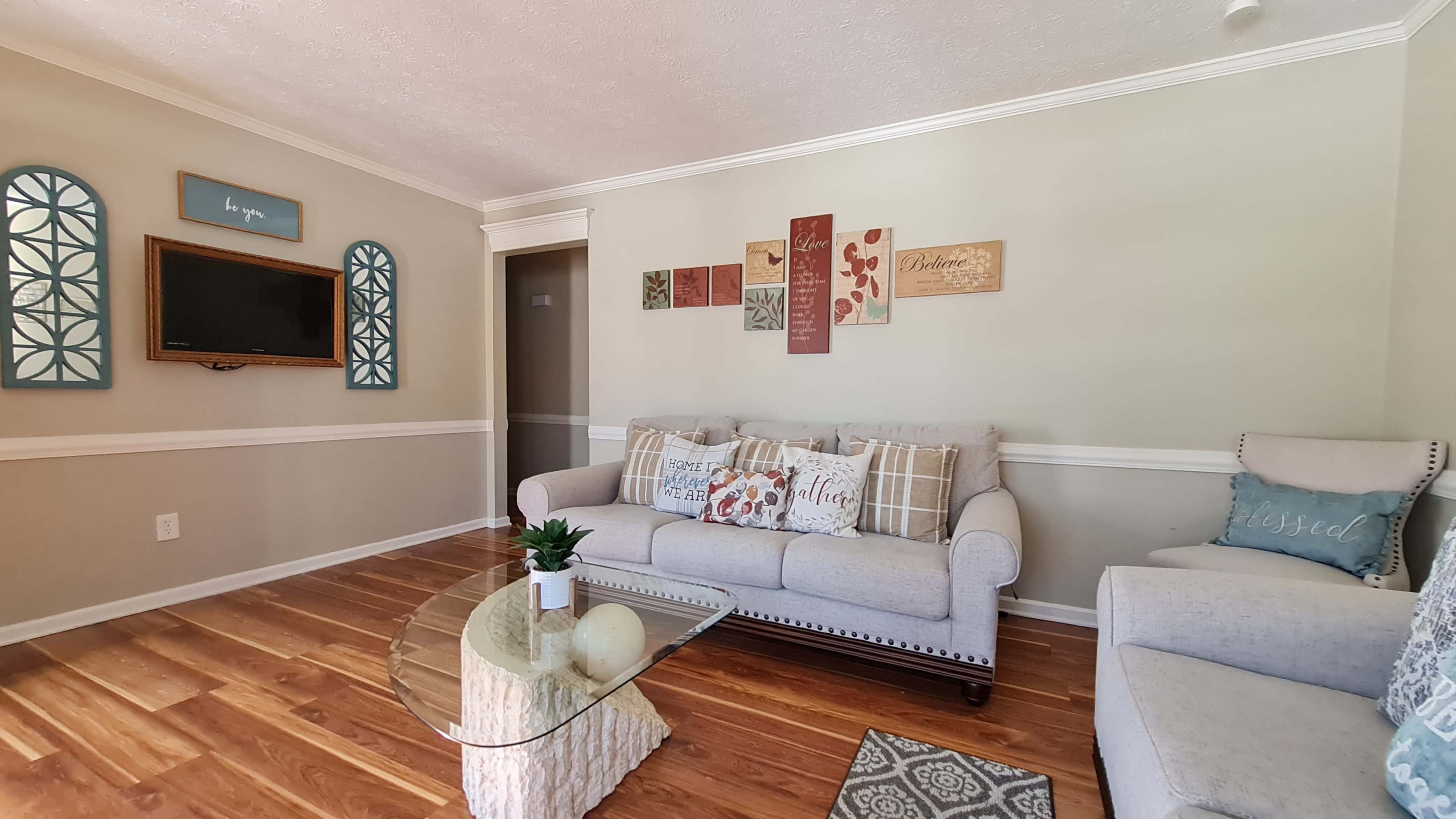 The living room features a light-colored sofa and a glass coffee table, with decorative wall art and neutral decor elements.