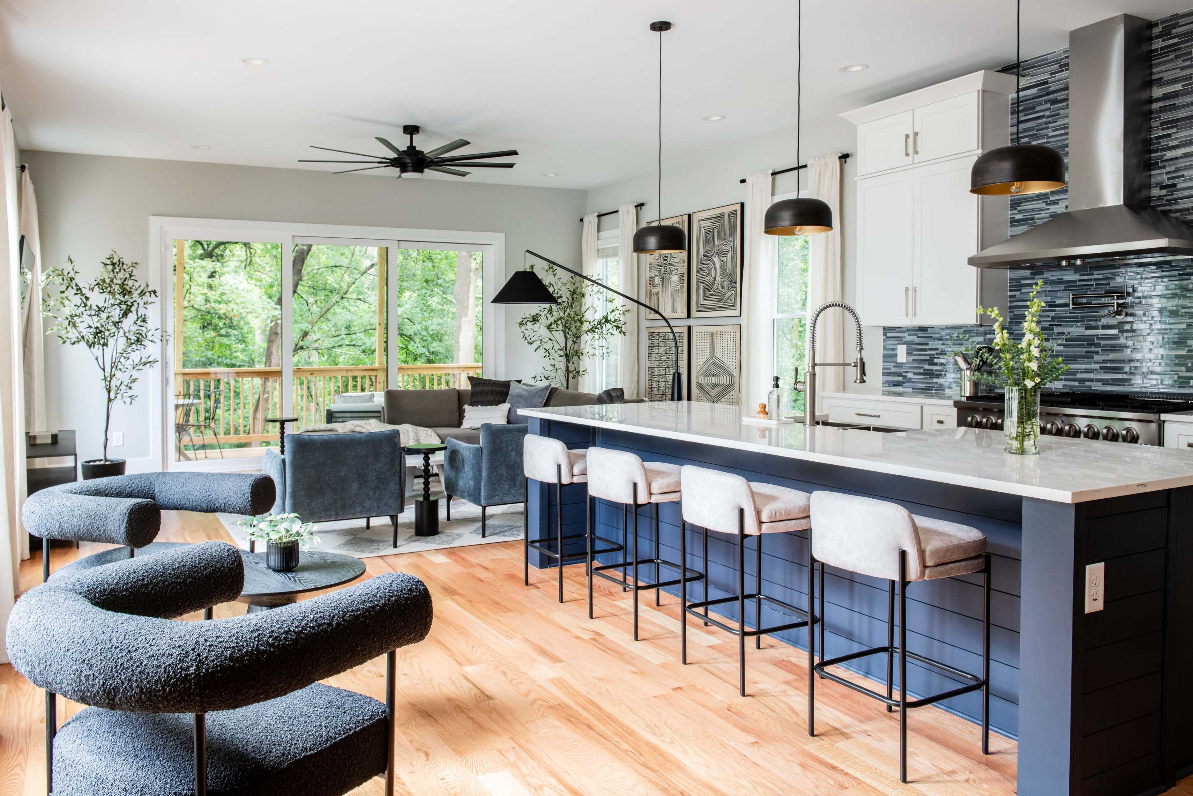A modern kitchen with a large island, bar stools, and a living area featuring comfortable seating and large windows overlooking a deck.
