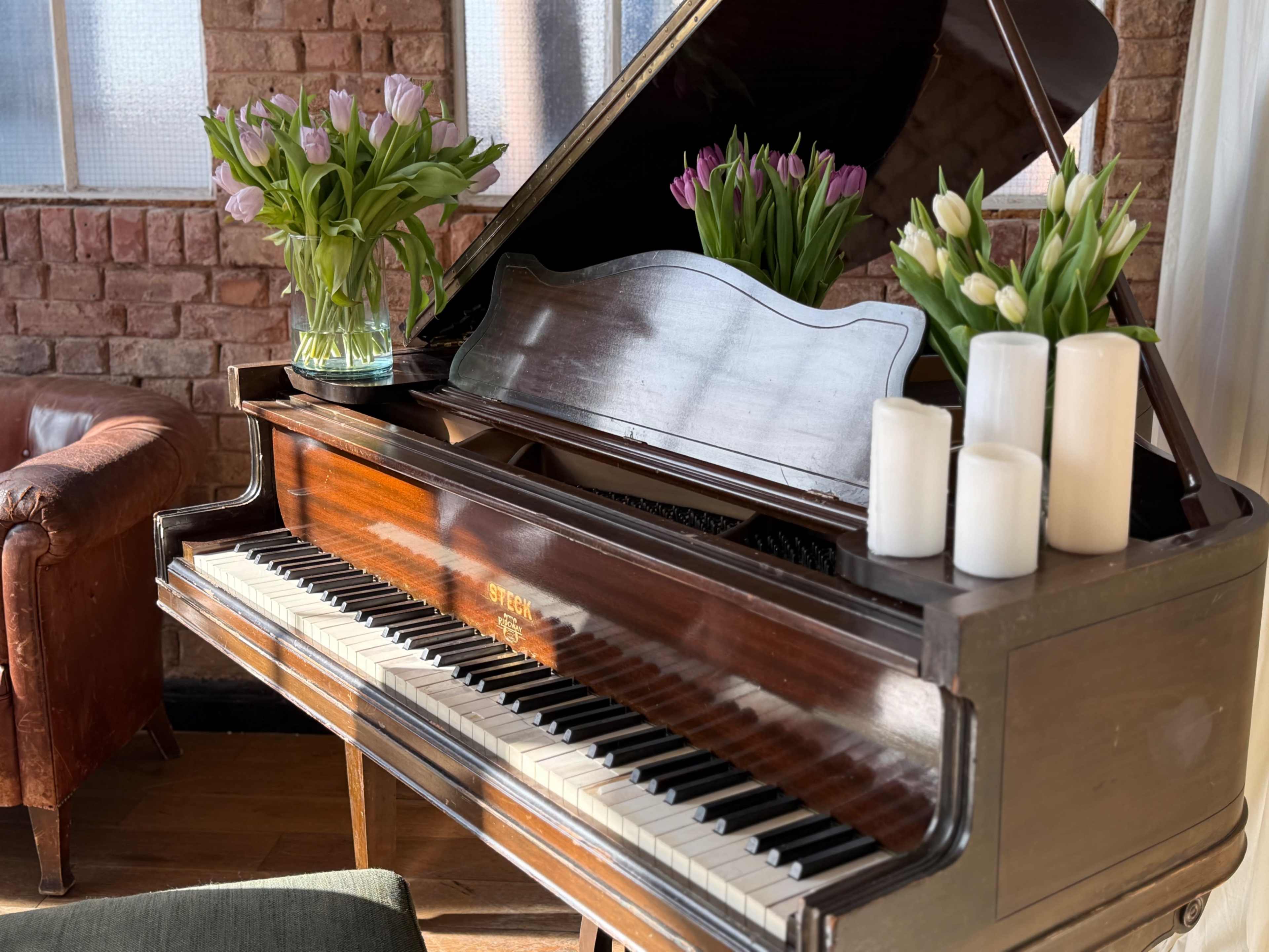 A polished grand piano is positioned in a room with brick walls, adorned with vases of tulips and candles on its surface.