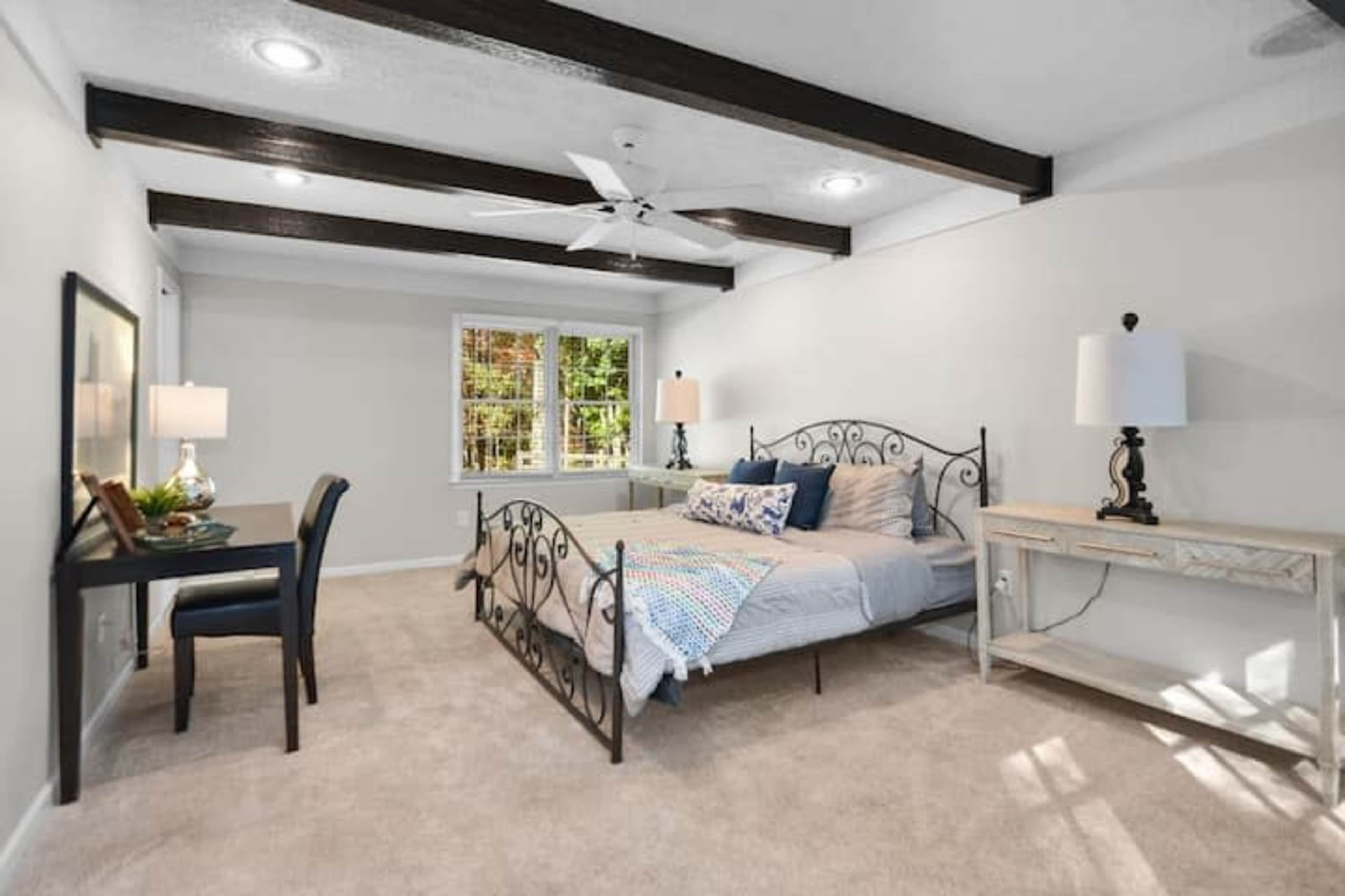 A bright bedroom with a metal bed frame, a desk set against the wall, and windows letting in natural light.
