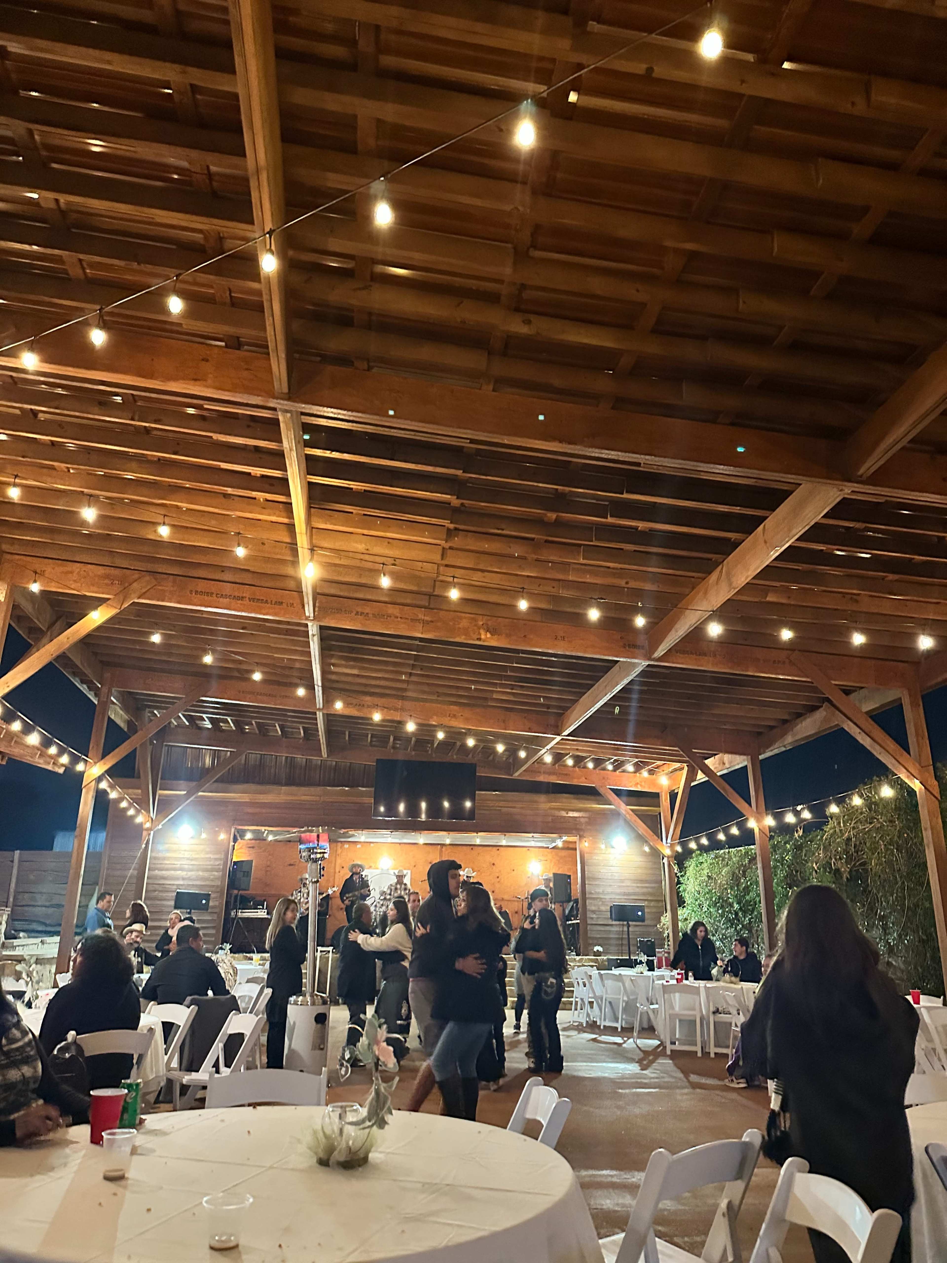 The image shows a large wooden pavilion with string lights overhead, where people are dancing and tables are set up for an event.