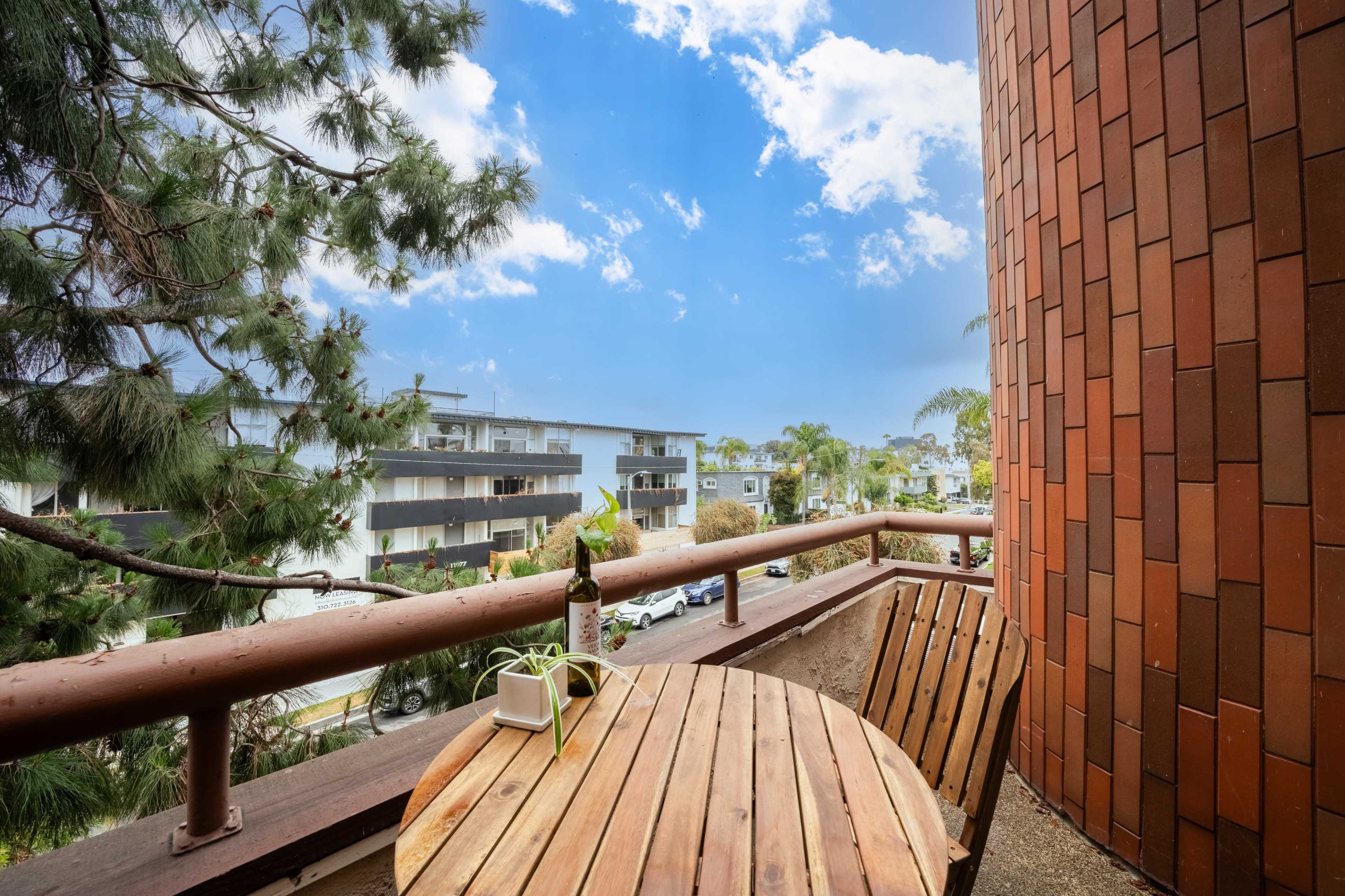 A wooden table and chair sit on a balcony with a view of surrounding buildings and palm trees under a partly cloudy sky.