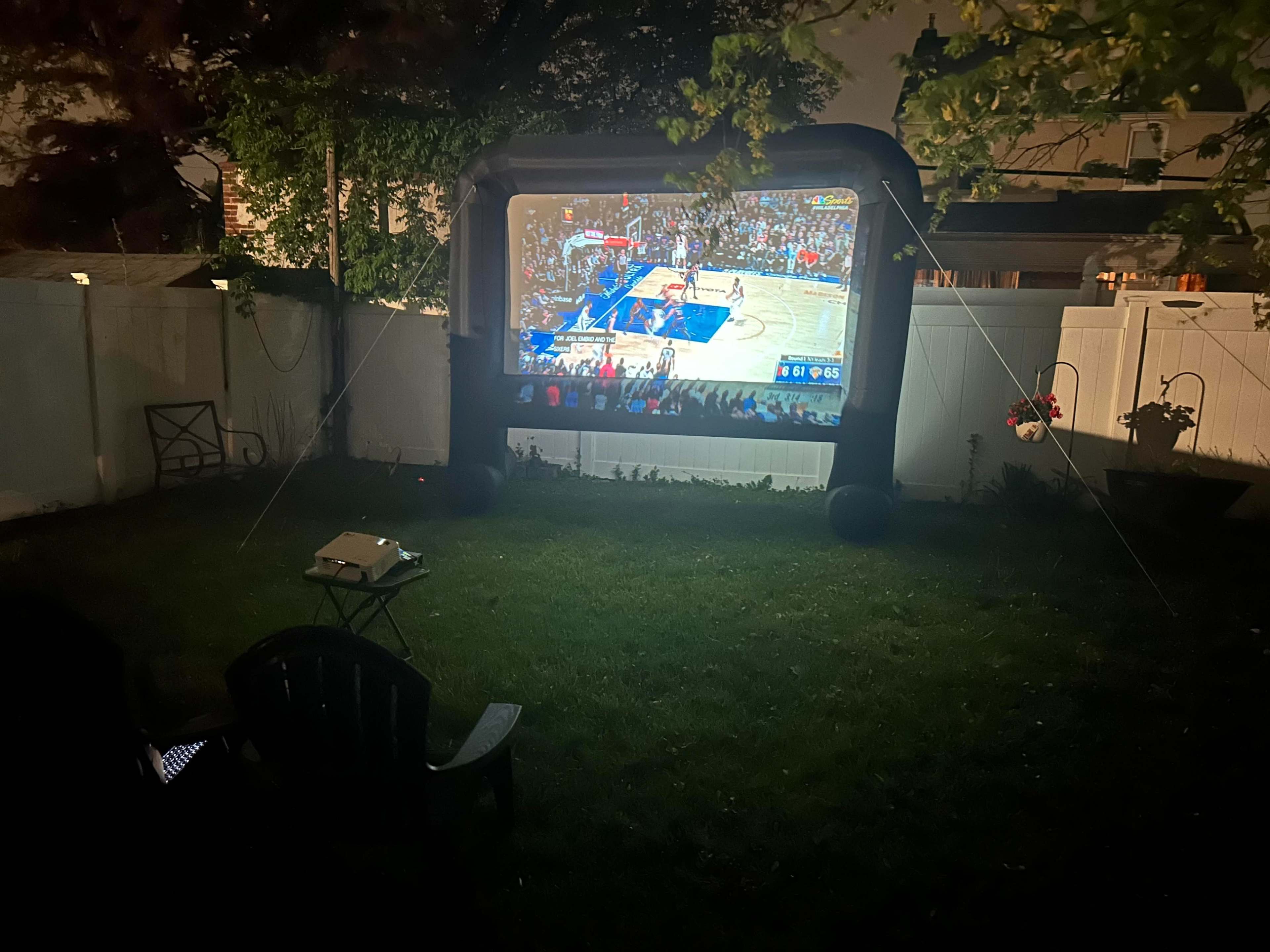 A large inflatable screen displays a basketball game in a backyard at night, with a projector set up beside it.