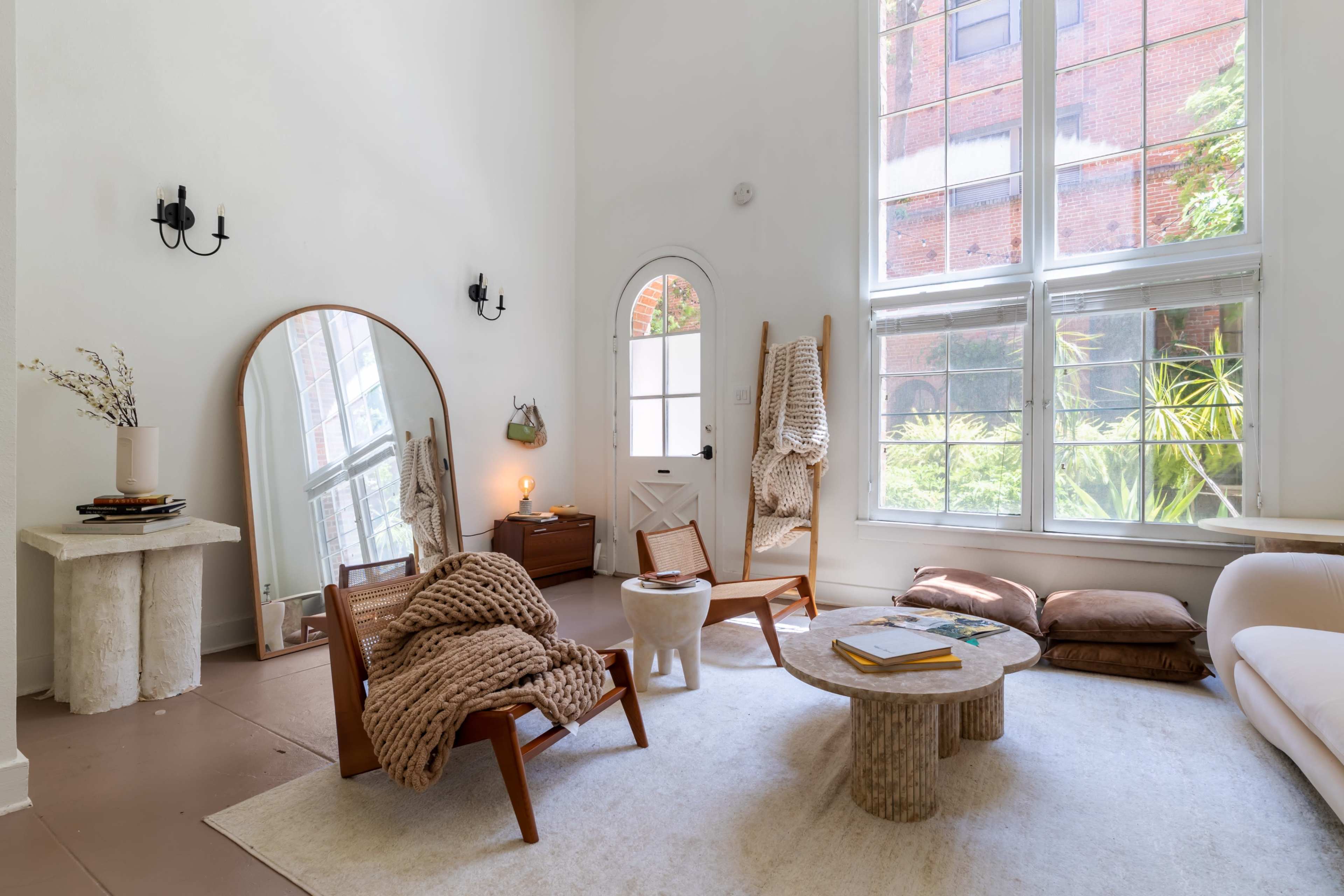 A bright, minimalist living room featuring wooden chairs, a round coffee table, a large mirror, and windows overlooking a garden.