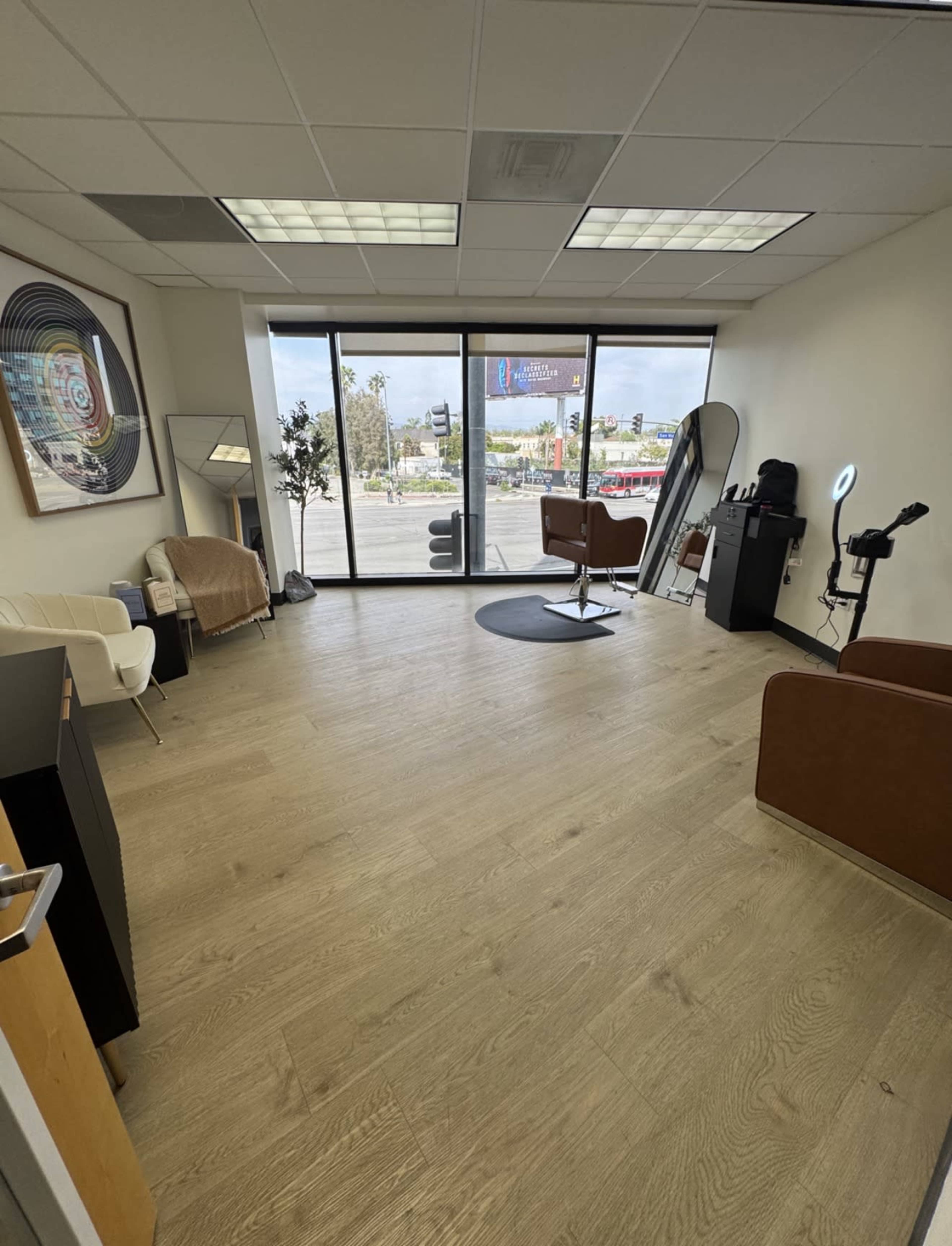 The image shows an interior office space with two brown chairs, a white couch, a large window displaying a view of the street, and minimalist decor.