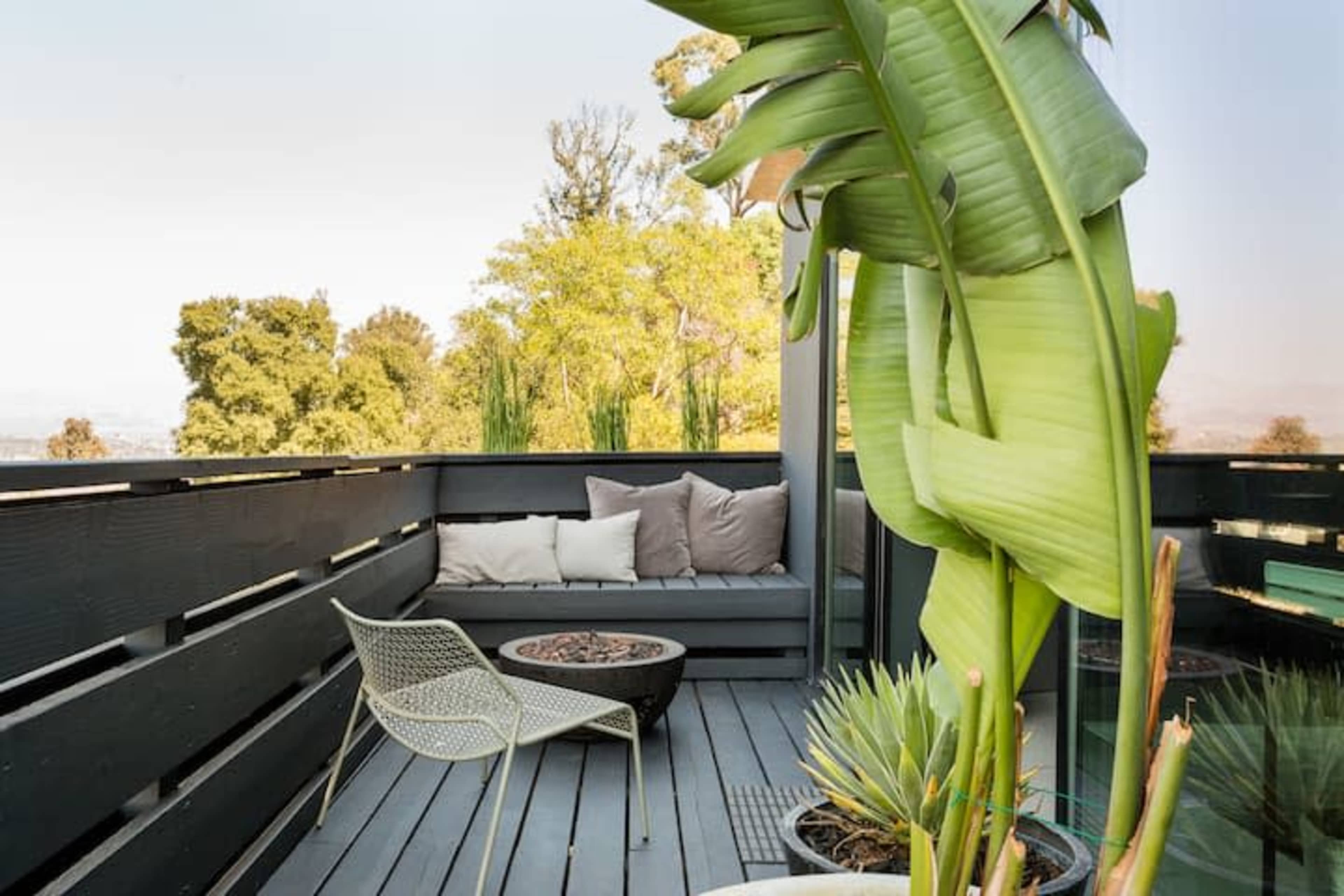 A cozy outdoor seating area features a low couch with cushions, a round coffee table, and large potted plants surrounded by wooden decking.