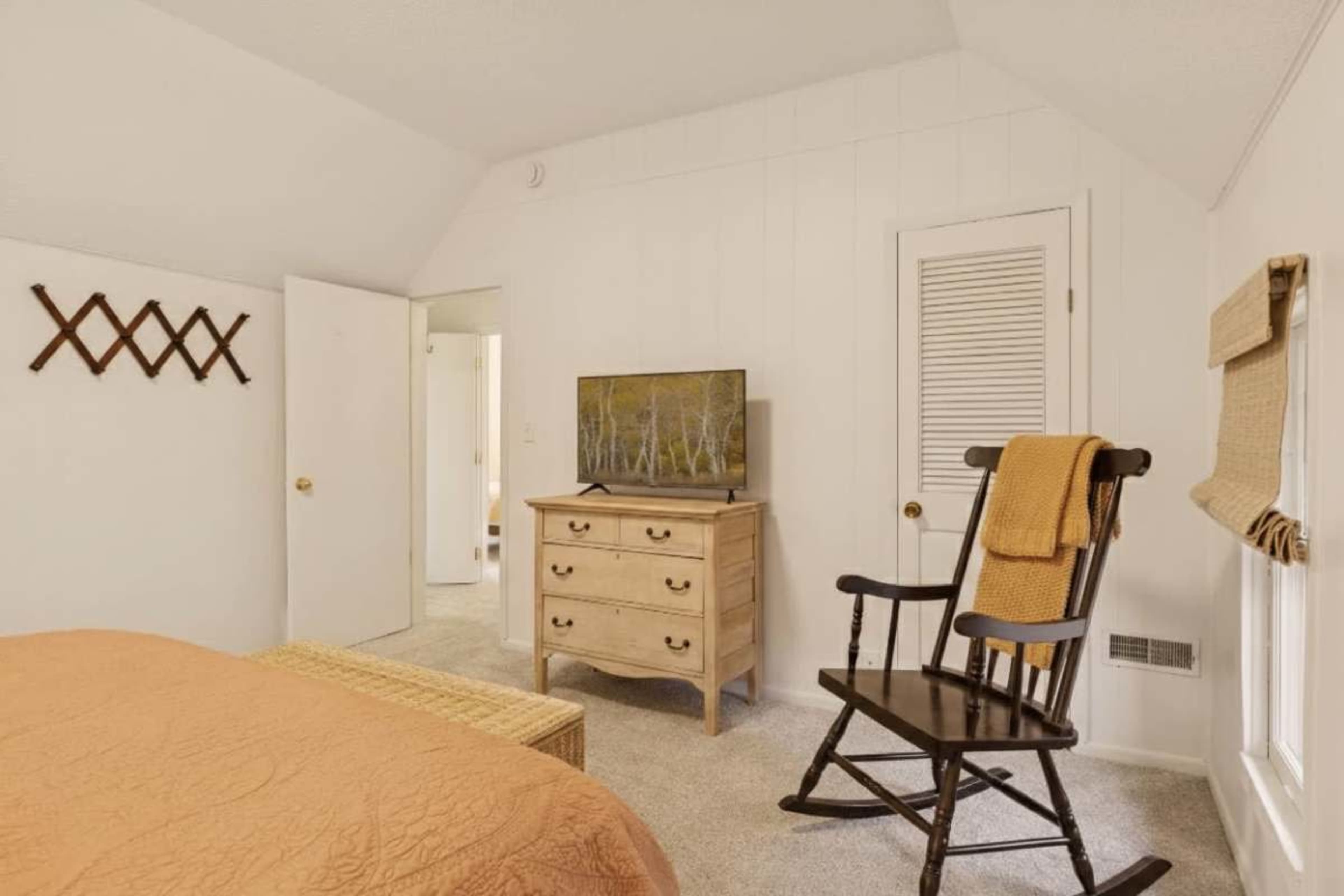 The room features a bed with an orange coverlet, a wooden dresser, a rocking chair, and a television placed on the dresser, all set against white walls.