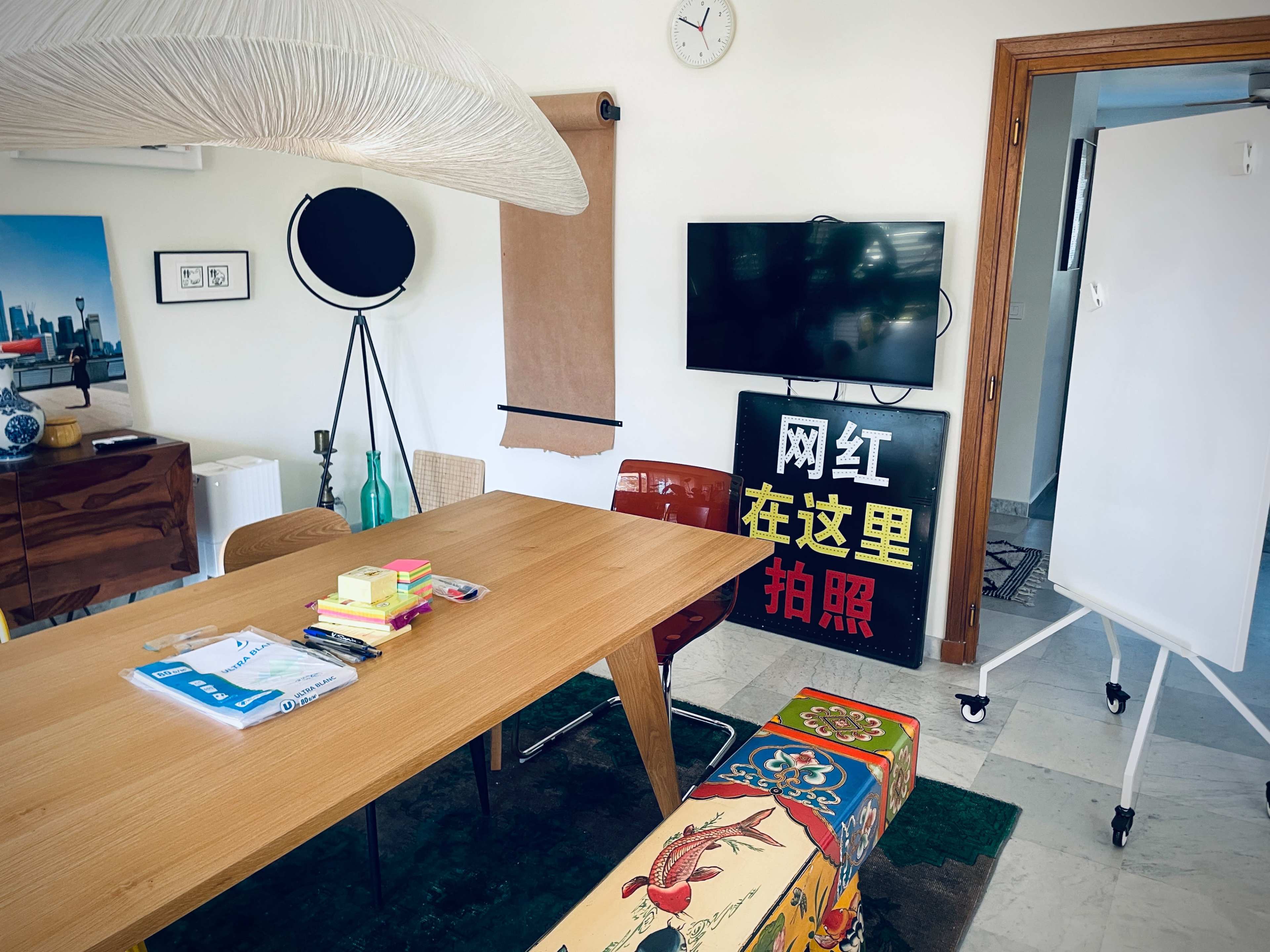 A bright meeting room features a wooden table surrounded by chairs, a large television, and a whiteboard, with decorative items and artwork on the walls.